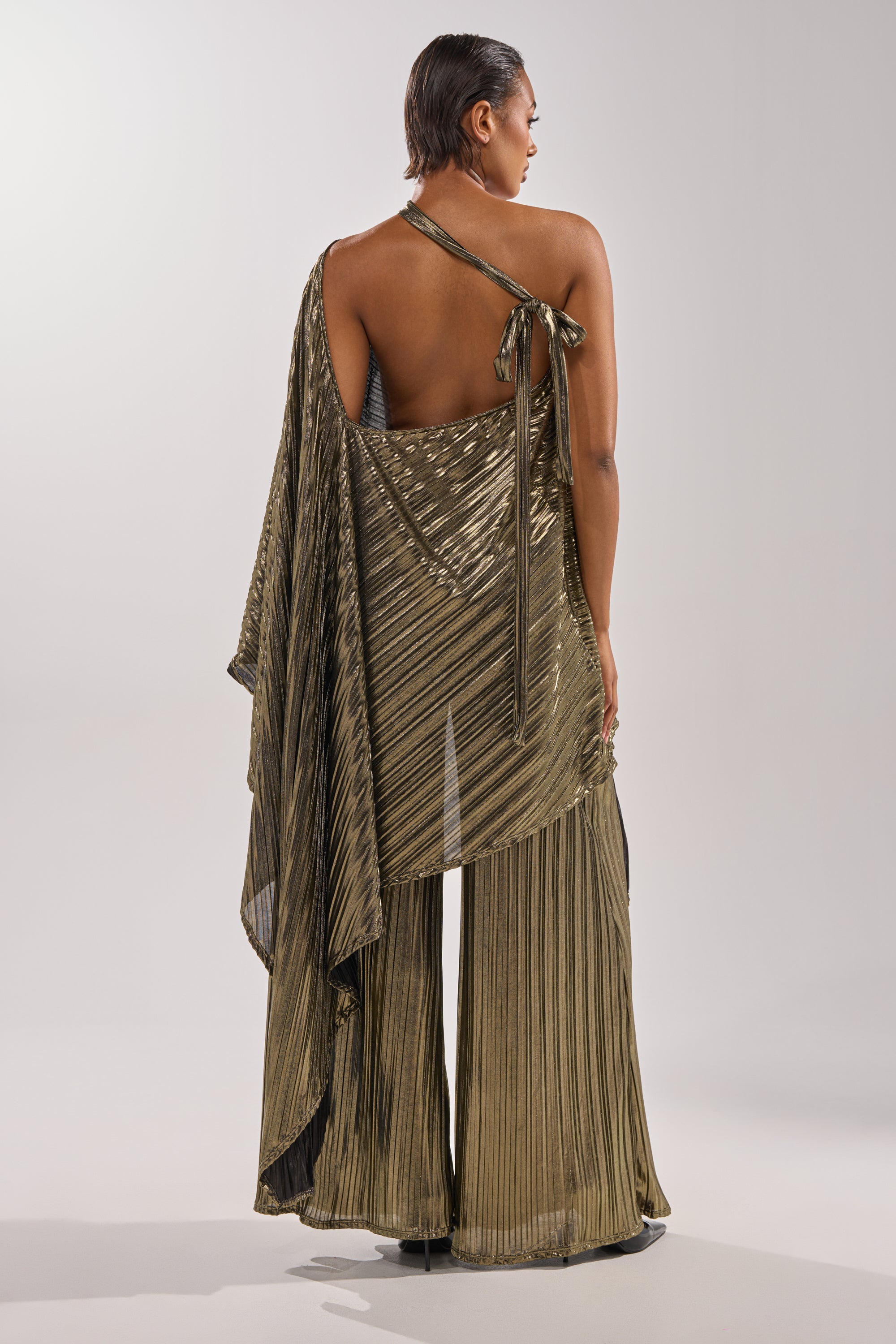 A person faces away from the camera, showcasing the BEST DRESSED BLOUSE—a striking metallic pleated halter top with an asymmetrical sash—paired with matching wide-leg pants, ideal for elegant evenings. Their hair is short and sleek. 9