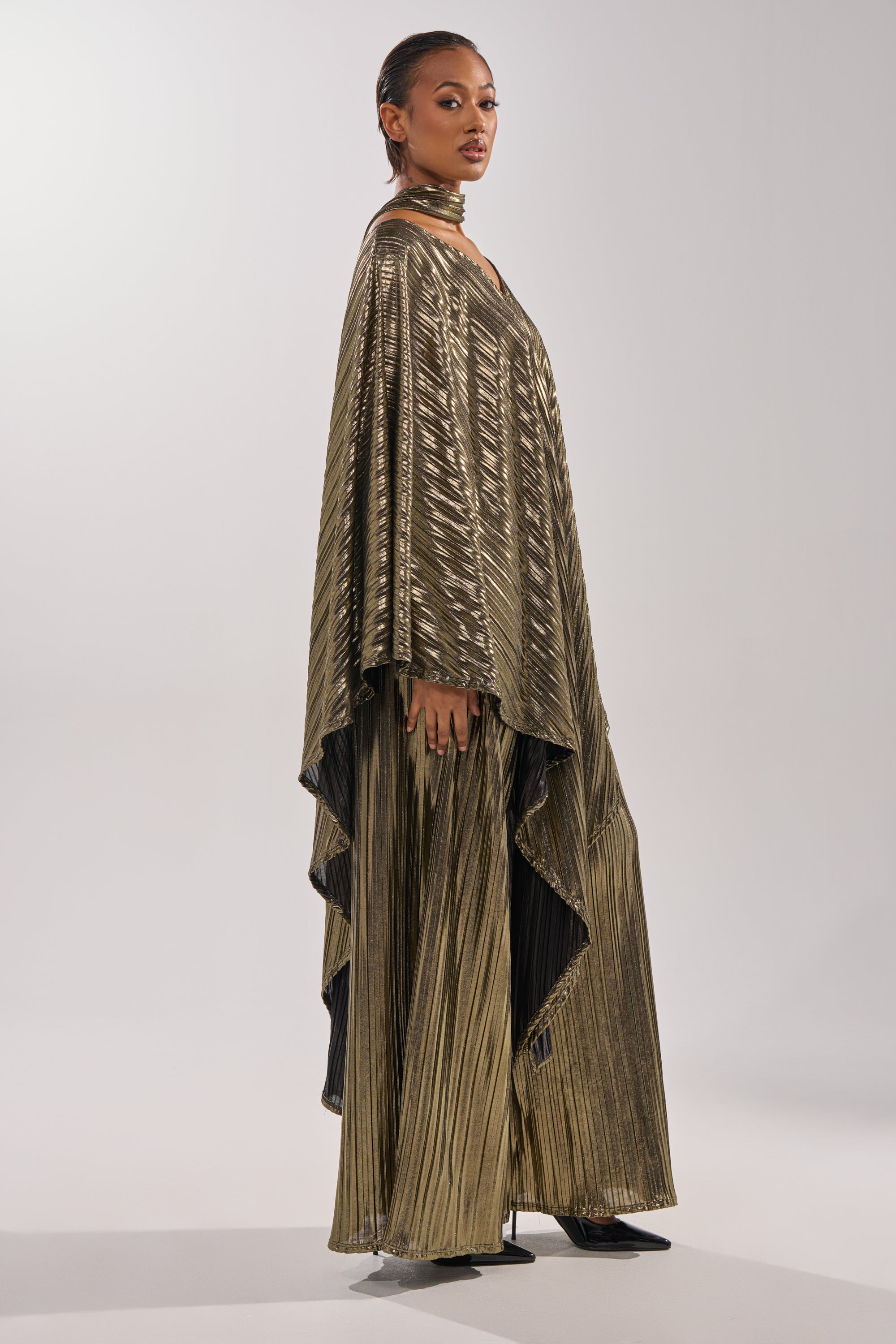 A woman models the BEST DRESSED BLOUSE—a gold metallic pleated top with wide sleeves—paired with matching pants and black pointed heels, set against a plain light background for an effortlessly sophisticated evening look. 11
