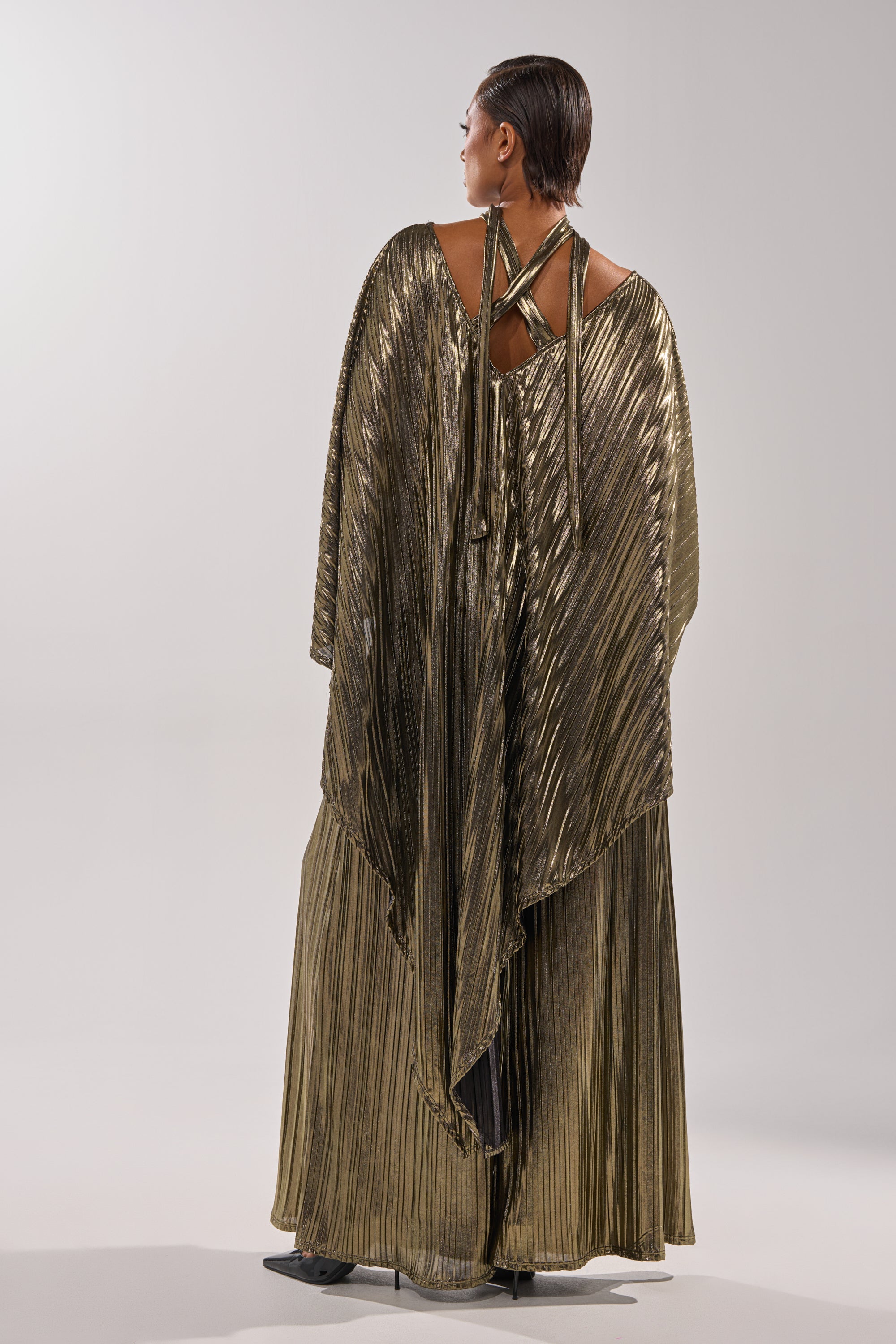 A person stands facing away, wearing the BEST DRESSED BLOUSE in metallic gold with pleats, a flowing cape, and crisscross back straps, blending elegance against a plain light background. 12