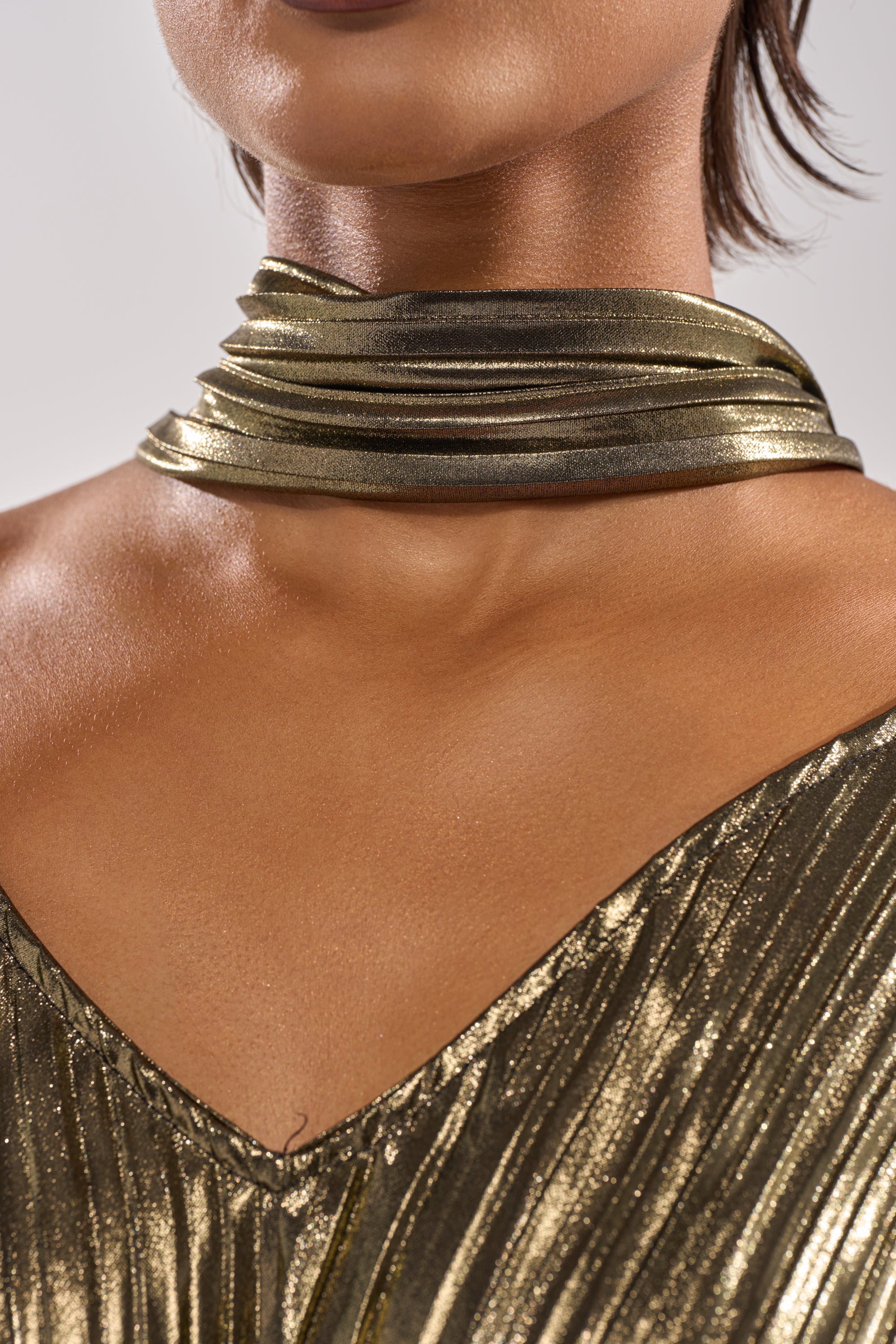 A close-up of someone wearing the BEST DRESSED BLOUSE, a shiny metallic pleated top paired with a matching gold scarf. The shot highlights the smooth, glowing skin on their neck and upper chest. 13