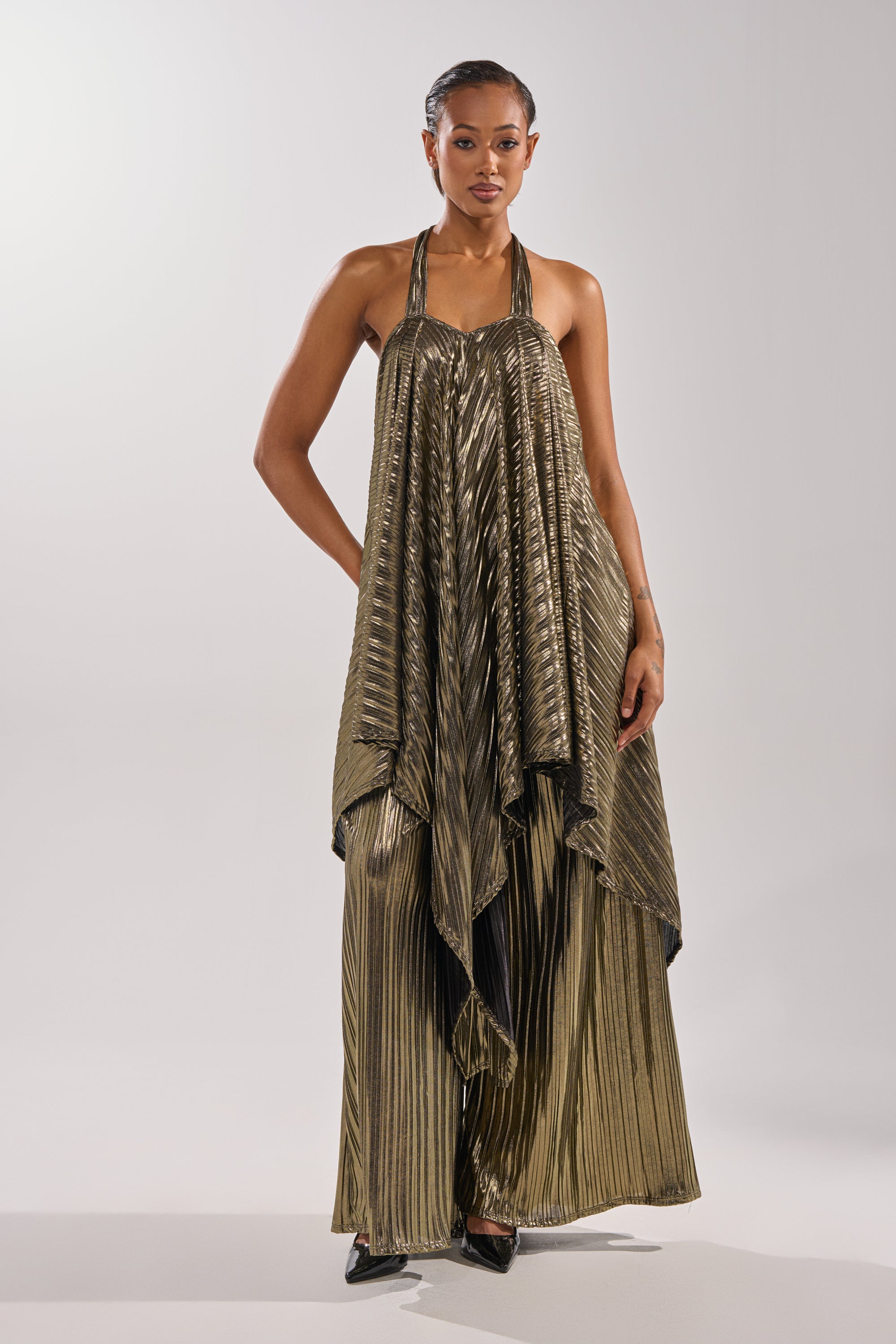 A woman wears the BEST DRESSED BLOUSE, a loose-fitting, shiny gold top with pleats and an asymmetrical hem, styled with black pointed shoes against a plain light background. 1