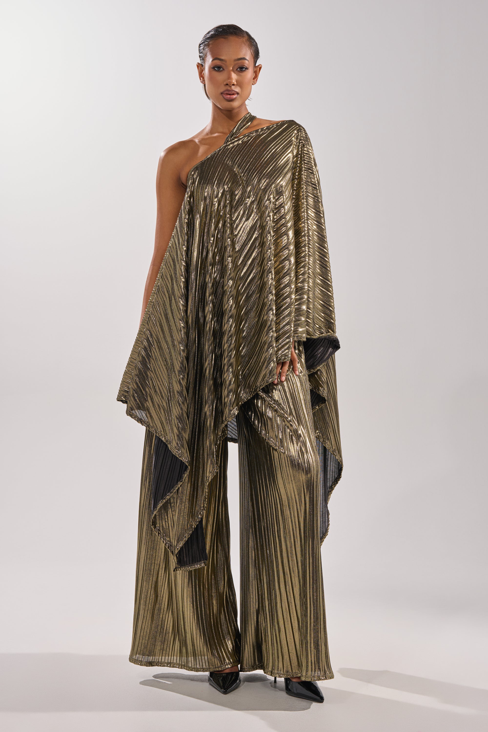 A woman poses in the BEST DRESSED BLOUSE, a shiny gold metallic pleated one-shoulder top, paired with matching wide-leg pants and black pointed-toe heels, standing against a plain light background. 2