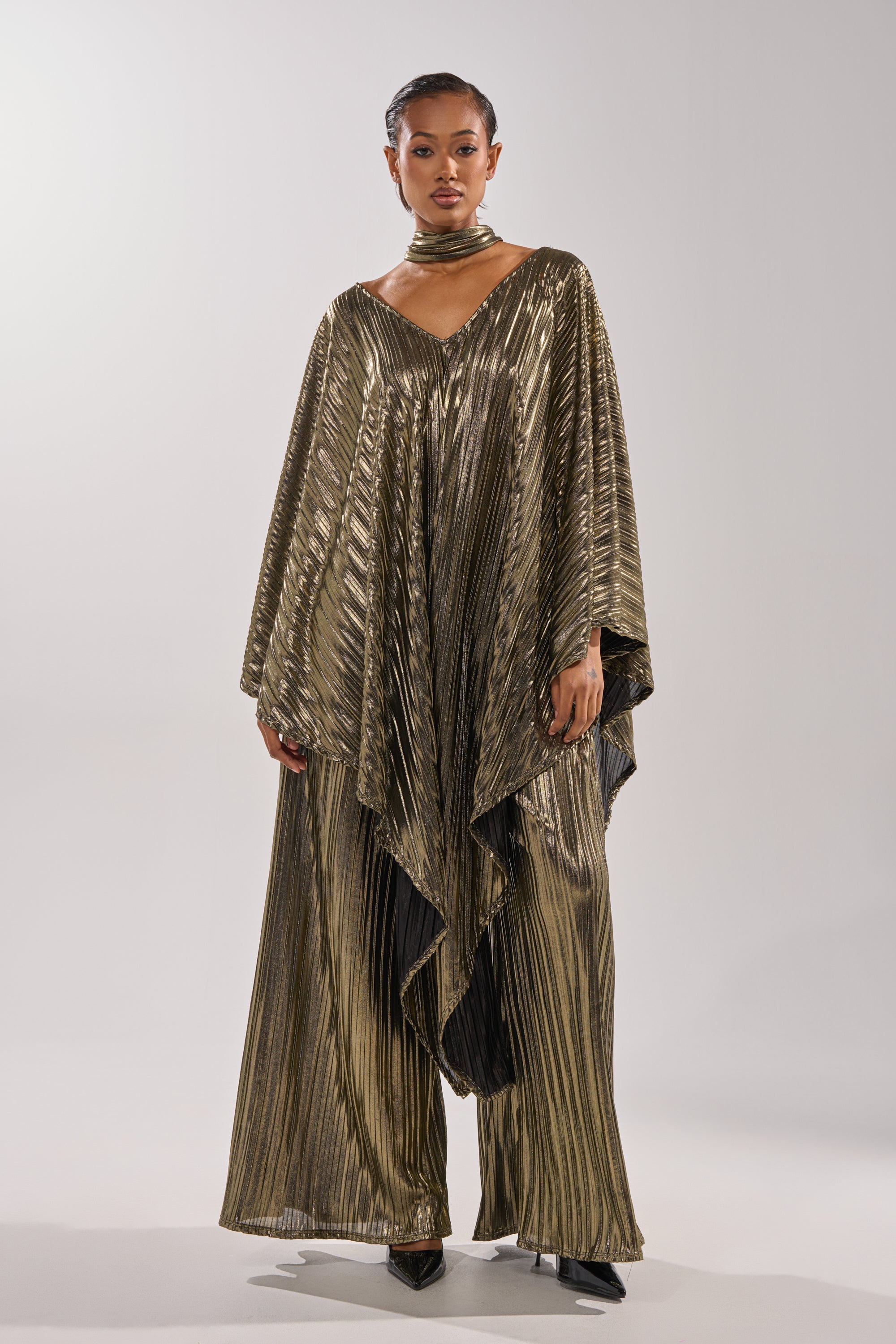 A woman models the BEST DRESSED BLOUSE, featuring shiny gold metallic pleats and wide kaftan sleeves, paired with wide-leg pants, a matching choker, and black pointed heels—ideal for standout evening style on a simple background. 3