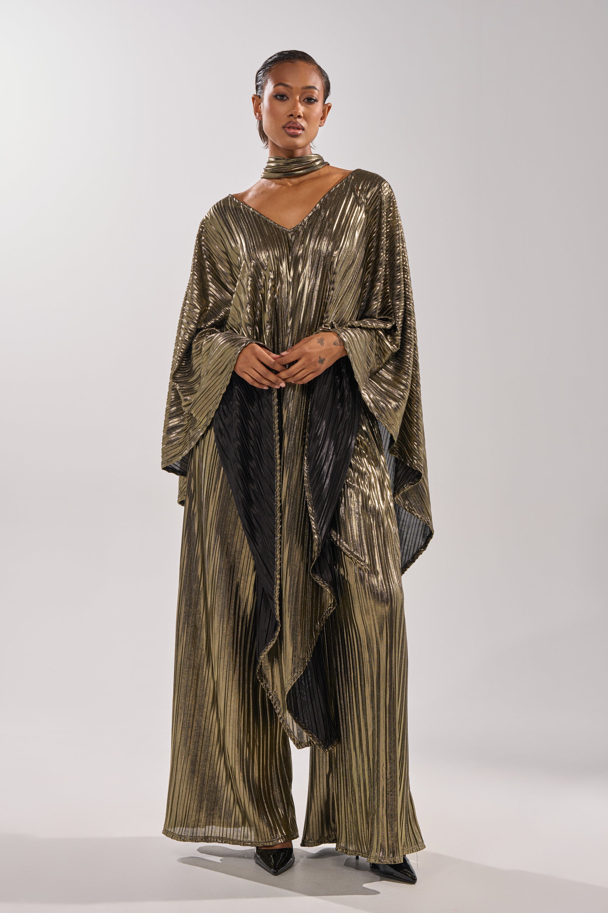 A woman wears the BEST DRESSED BLOUSE, a metallic pleated top with cape silhouette and wide sleeves, paired with matching pants, a choker, and black pointed heels against a plain light background. 4