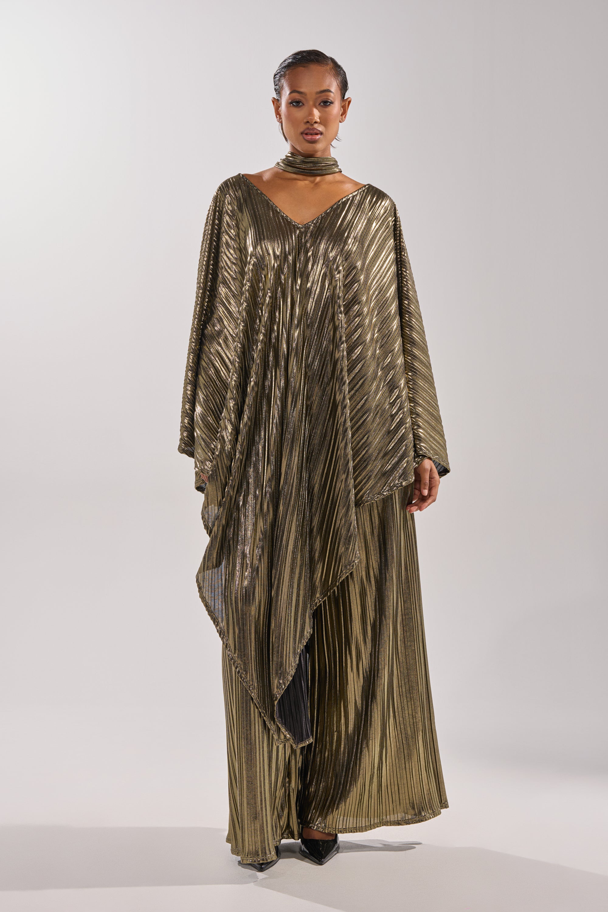 A woman faces forward wearing the BEST DRESSED BLOUSE, a flowing metallic gold top with wide sleeves, pleats, and a deep V neckline. She poses against a plain light background in this kaftan-inspired evening look. 5