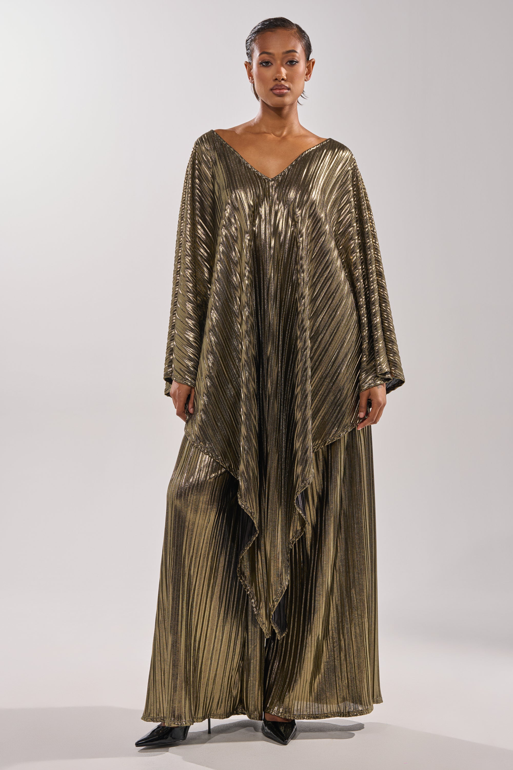 A woman faces forward in the BEST DRESSED BLOUSE, a flowing, metallic gold pleated top with wide sleeves and an asymmetric hem. She pairs it with black pointed boots and stands against a plain light background. 6