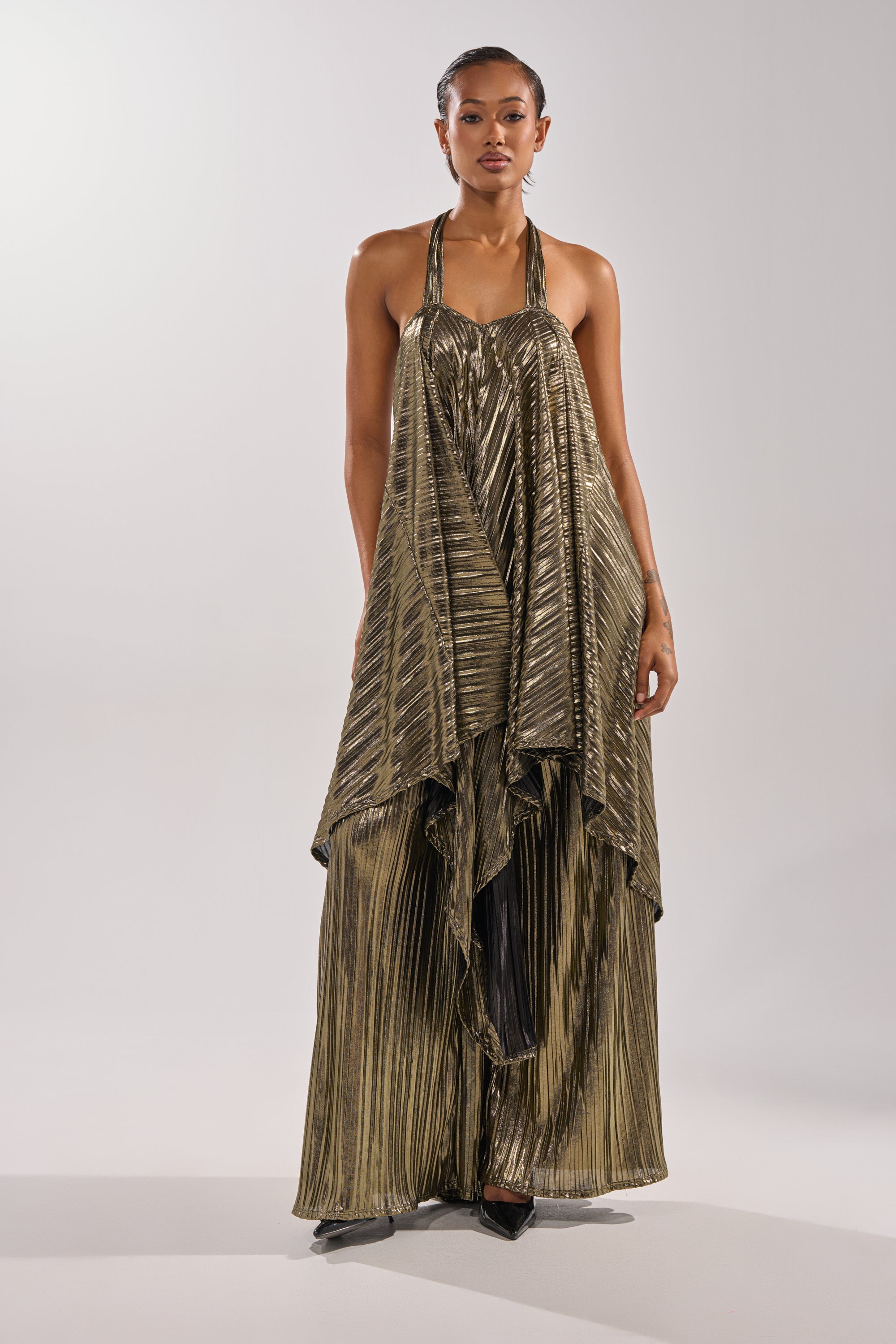 A woman stands facing forward in the BEST DRESSED BLOUSE, featuring flowing metallic gold and black stripes with a halter neckline and asymmetrical hem. She has slicked-back hair, neutral makeup, and poses against a plain light background. 7