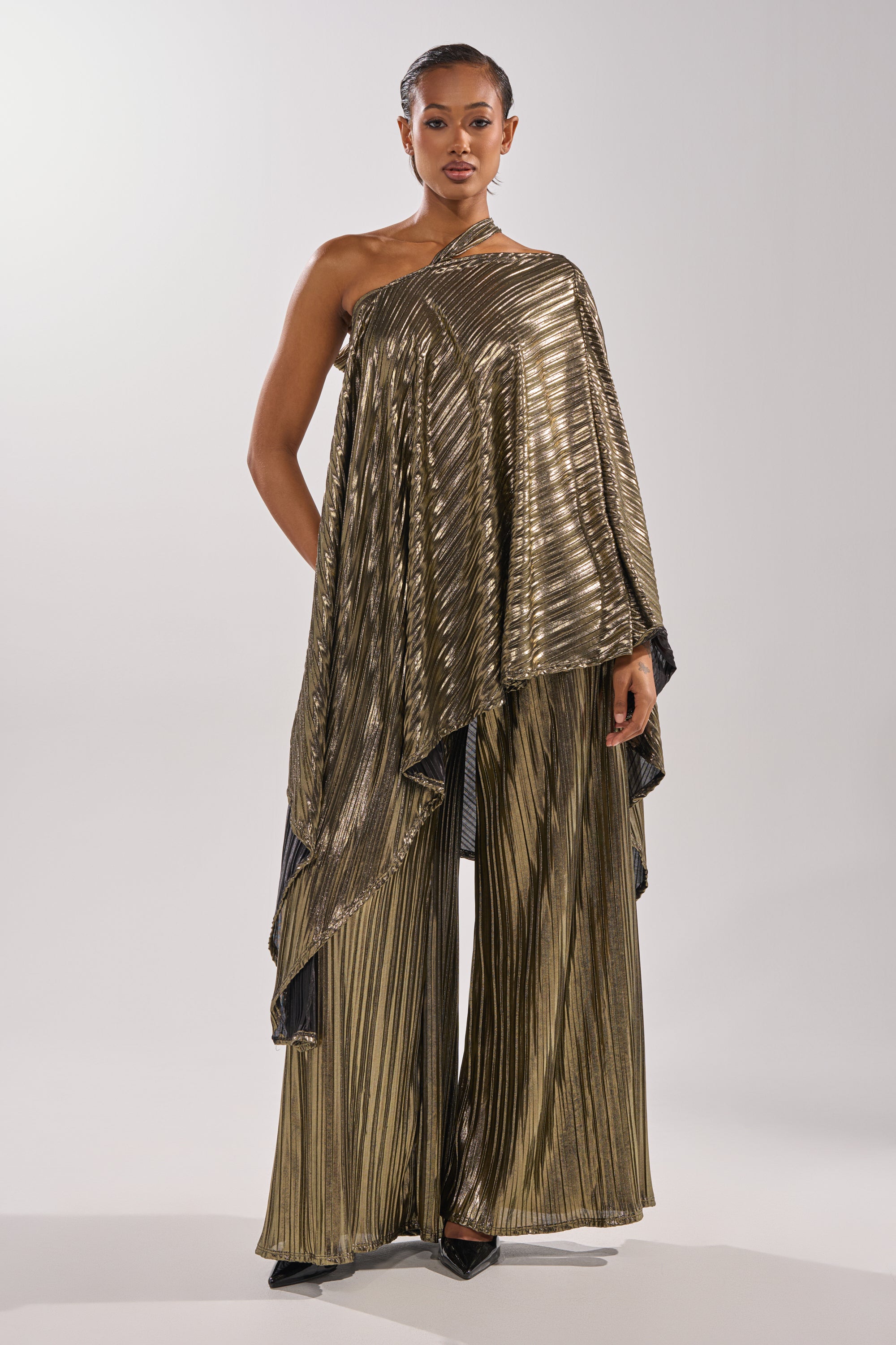 A person stands confidently in the BEST DRESSED BLOUSE—a metallic pleated top with matching wide-leg pants and black pointed shoes—showcasing chic modern evening wear against a plain light background. 8