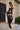 A woman poses outdoors in the SEE YOU LATER LACE BLOUSE styled over black lingerie, thigh-high stockings, boots, and a black and white cap. She stands by a building with one leg bent near a gate and greenery.