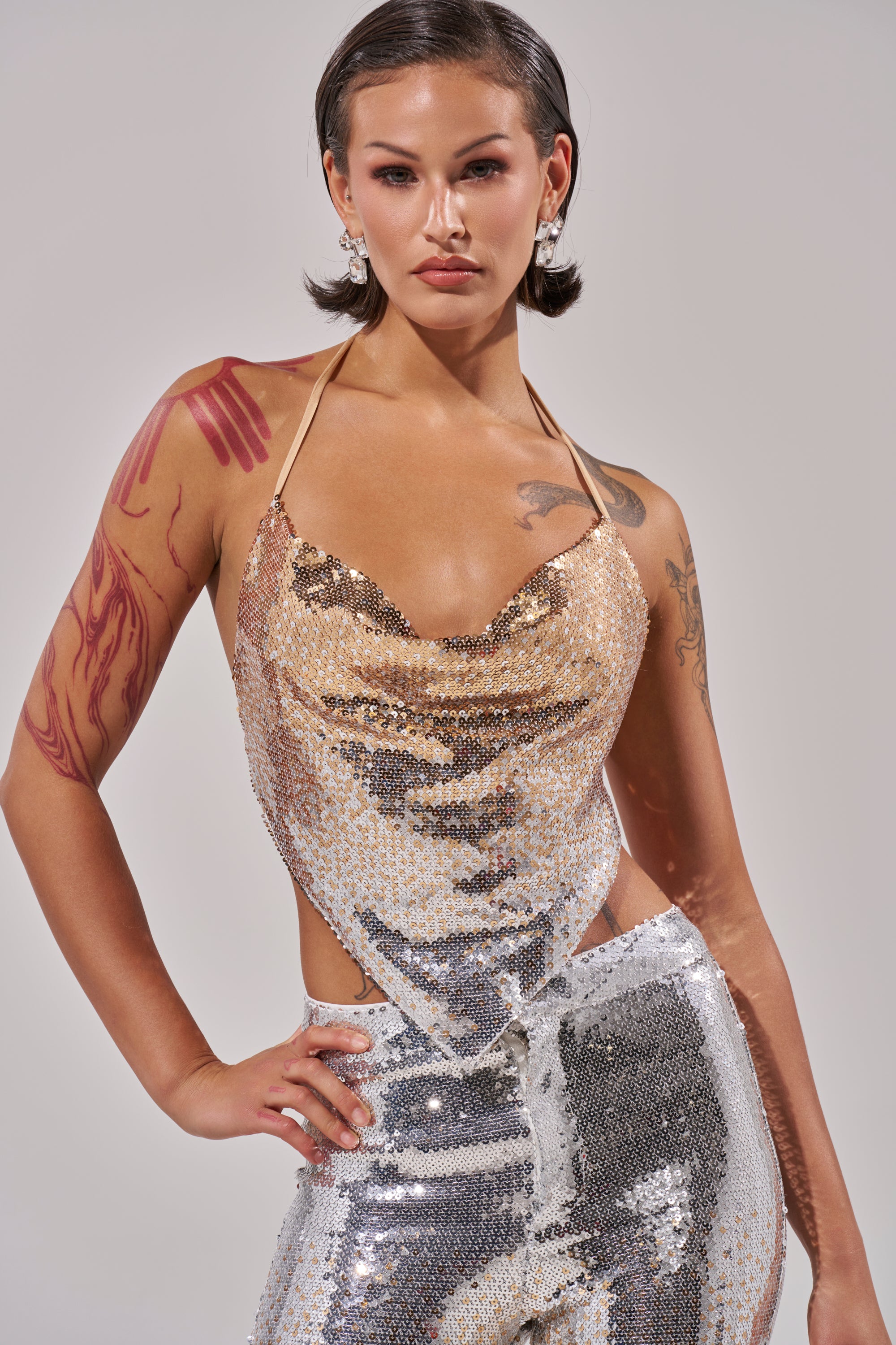 A person with short dark hair and visible arm tattoos confidently models the GALAXY OMBRE SEQUIN HALTER TOP with matching pants, maintaining a neutral facial expression.