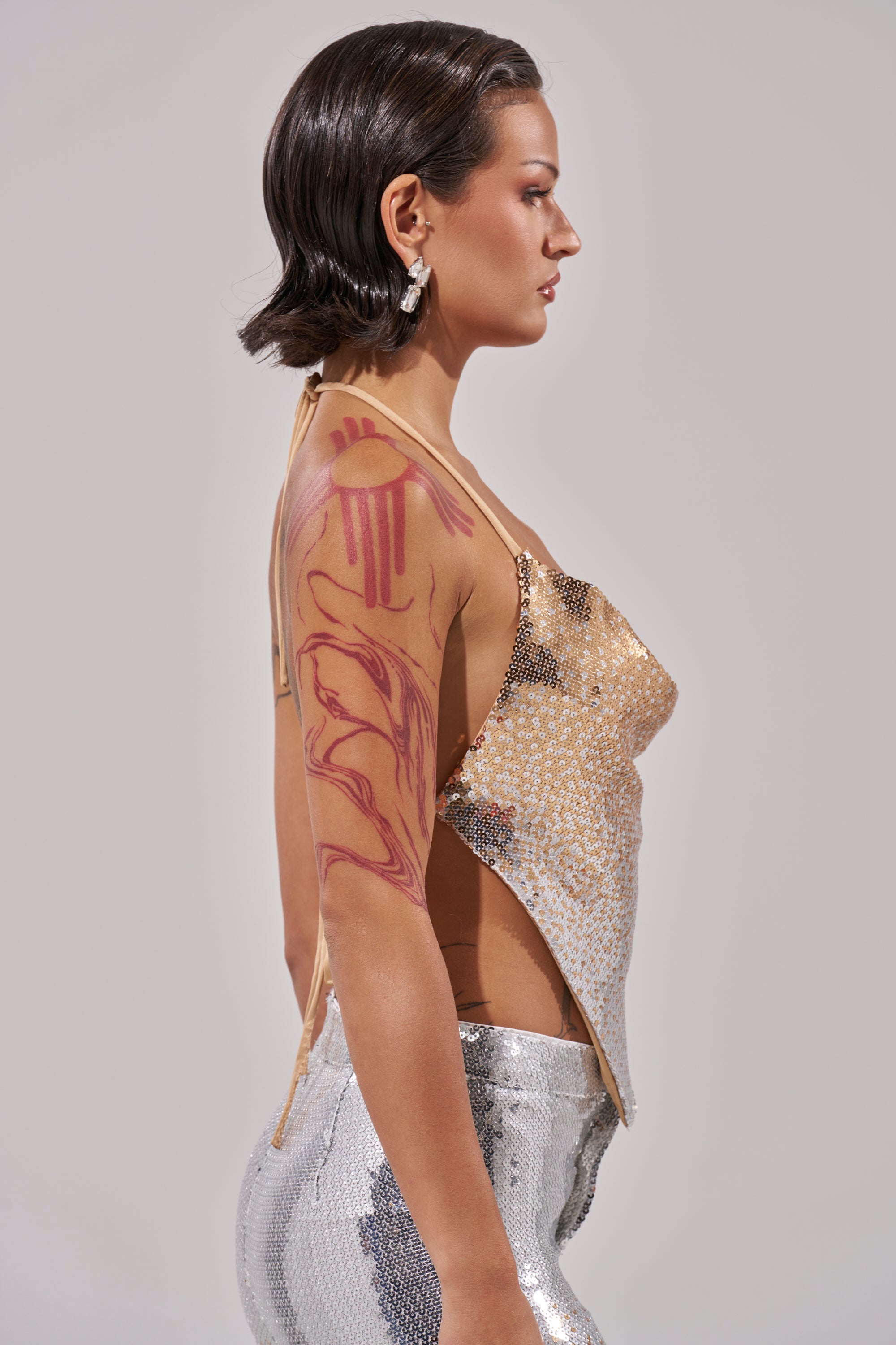 A woman with short dark hair, wearing shiny silver pants and the GALAXY OMBRE SEQUIN HALTER TOP, stands in profile. She has a bold red abstract upper arm tattoo, statement earrings, and is set against a neutral gray background.