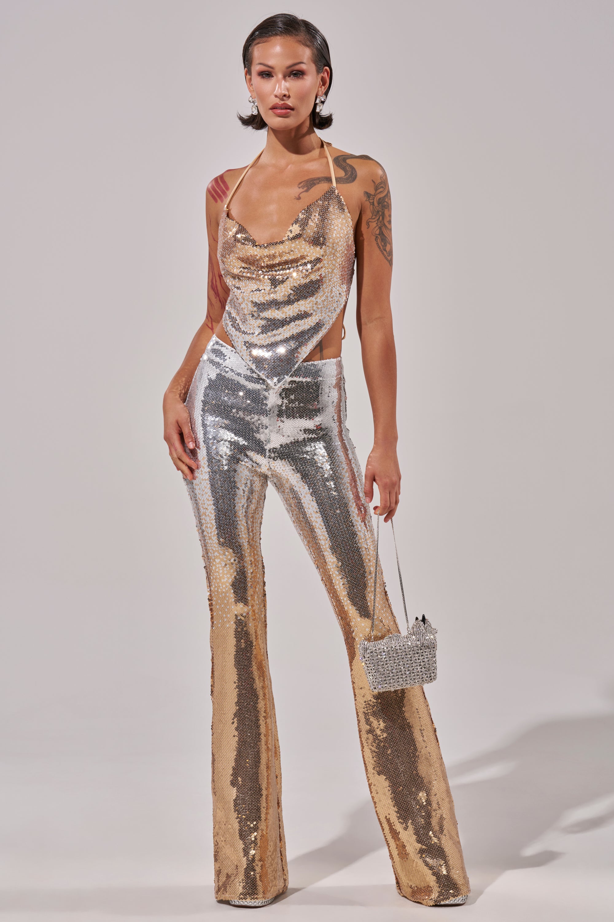 A woman with tattoos and short, styled hair poses confidently in the GALAXY OMBRE SEQUIN HALTER TOP and matching flared pants, holding a sparkling handbag against a plain background.