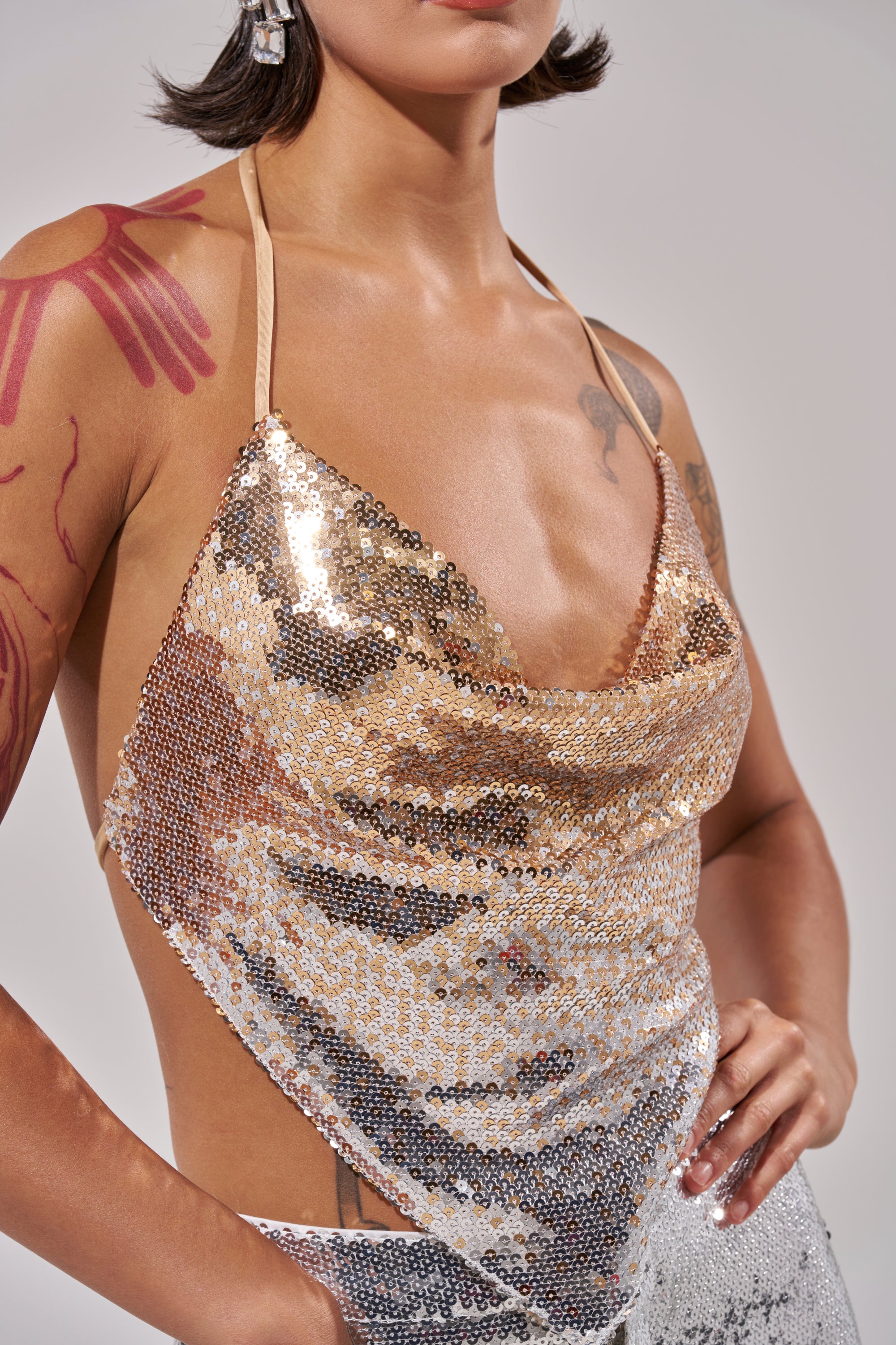 A person wearing the GALAXY OMBRE SEQUIN HALTER TOP poses with one hand on their hip, showing tattoos on their arm and chest against a neutral background. Their head is partially out of frame.