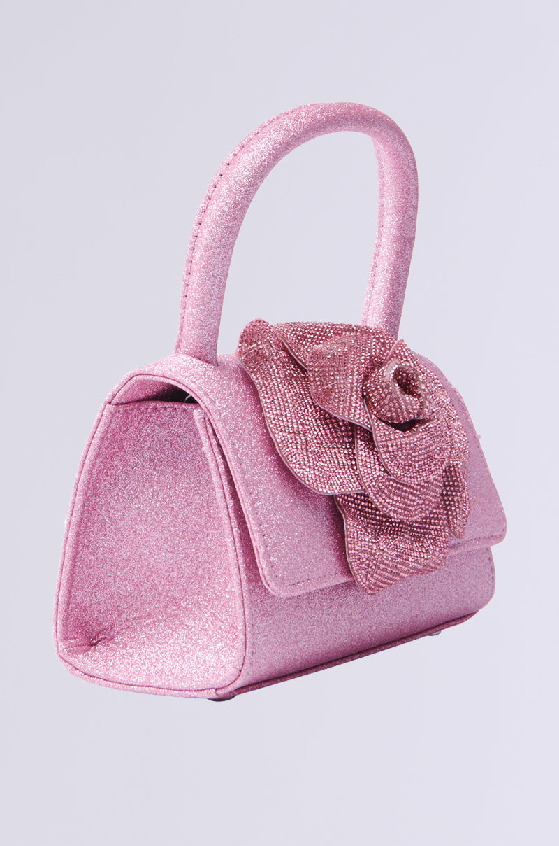 The DANCE ALL NIGHT FLORAL MINI BAG is a pink glitter handbag adorned with rhinestones and a large decorative rose. Featuring a curved handle and smooth, structured shape, it offers an elegant crossbody fit for any occasion.