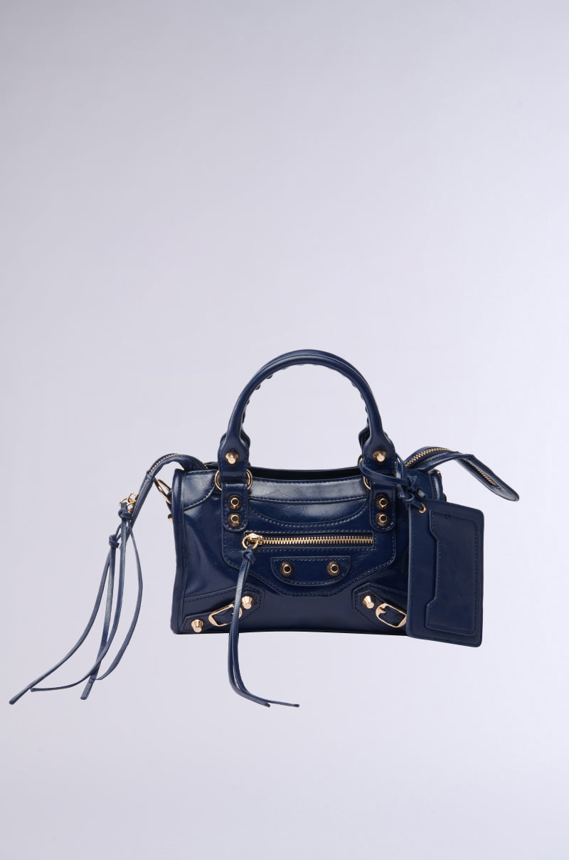 The IN YA CITY PURSE is a small, glossy navy faux leather bag by AKIRA Label, featuring gold hardware, zipper details, a top handle, stud accents, and hanging tassels, set against a plain light gray background. 1
