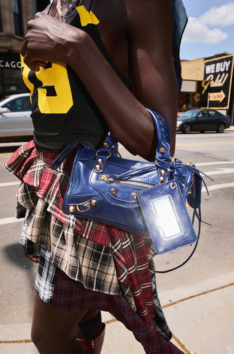 Walking down a city street, a person in a black sports jersey and plaid skirt carries the IN YA CITY PURSE—a navy faux leather crossbody bag. A “We Buy Gold” sign appears in the background. 0