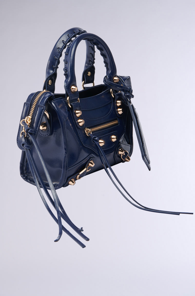 The IN YA CITY PURSE is a small, glossy navy faux leather bag from AKIRA Label with gold hardware, zipper accents, stud details, braided top handles, and long leather tassels on the zippers. 2