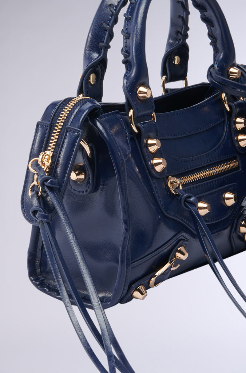 A close-up of the glossy navy IN YA CITY PURSE by AKIRA Label, featuring gold hardware, stud accents, zippers, buckles, double handles, and long decorative faux leather tassels. 3