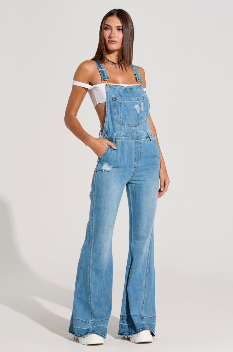 A woman poses in the LARINA DENIM OVERALLS, featuring medium wash denim with flared legs and distressed detailing. She pairs them with a white off-the-shoulder top and stands against a plain background, wearing white platform shoes. Her straight, shoulder-length hair frames her face as she faces forward with her hands in the front pockets.