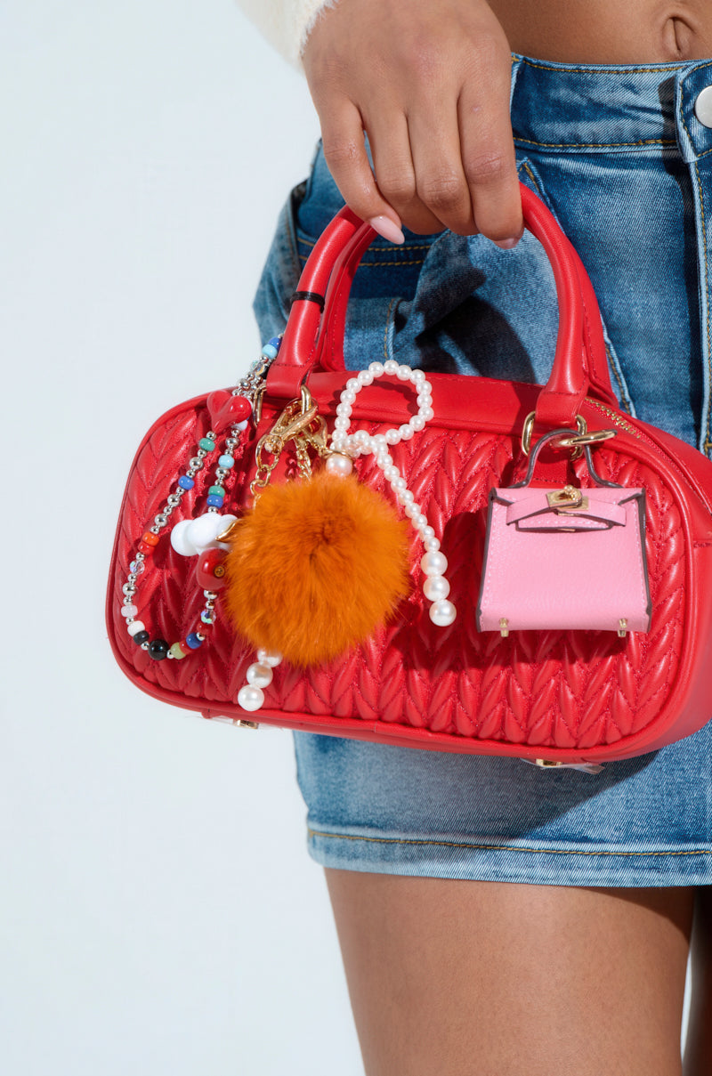 A person showcases the TOO TRENDY JUNK BAG, a cherry red quilted faux leather handbag with colorful beads, an orange furry keychain, and a pink purse charm. It features gold hardware and a detachable strap. They pair it with a denim skirt and white top against a plain backdrop.