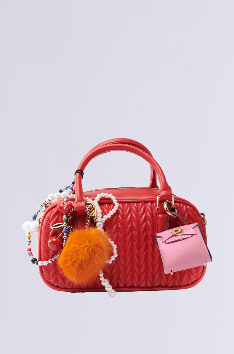 The TOO TRENDY JUNK BAG is a small cherry red faux leather quilted handbag with a short handle and detachable shoulder strap, adorned with decorative charms—a fluffy orange pom-pom, pink mini purse charm, and colorful bead string with pearls—against a plain gray background.