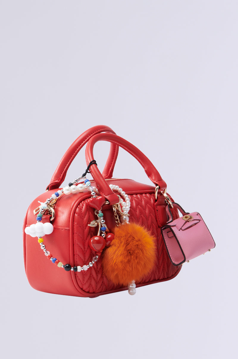 The TOO TRENDY JUNK BAG is a cherry red faux leather quilted handbag featuring charms, metallic gold hardware, beads, an orange pom-pom, and a small pink purse. It has a structured shape with two handles and a detachable shoulder strap against a plain background.