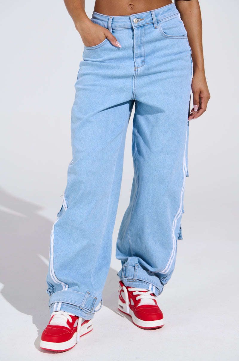 A person stands against a plain light background wearing LEAVE IT ON THE TRACK RELAXED LEG DENIM PANTS in light blue with white side stripes, cuffed hems, red and white sneakers, and one hand in a pocket.