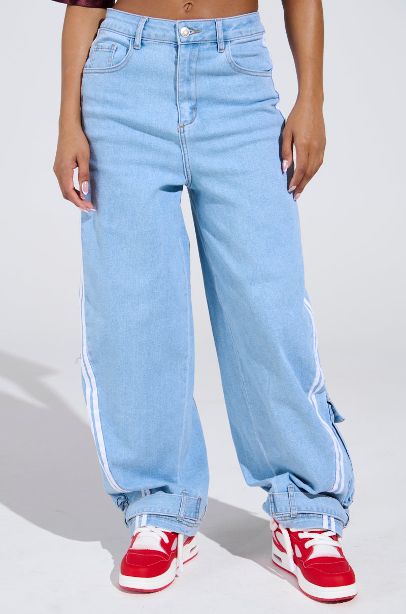 A person wearing LEAVE IT ON THE TRACK RELAXED LEG DENIM PANTS in light blue with white side stripes and a relaxed fit, paired with red-and-white sneakers, stands against a plain light background. Only the lower torso and legs are visible.