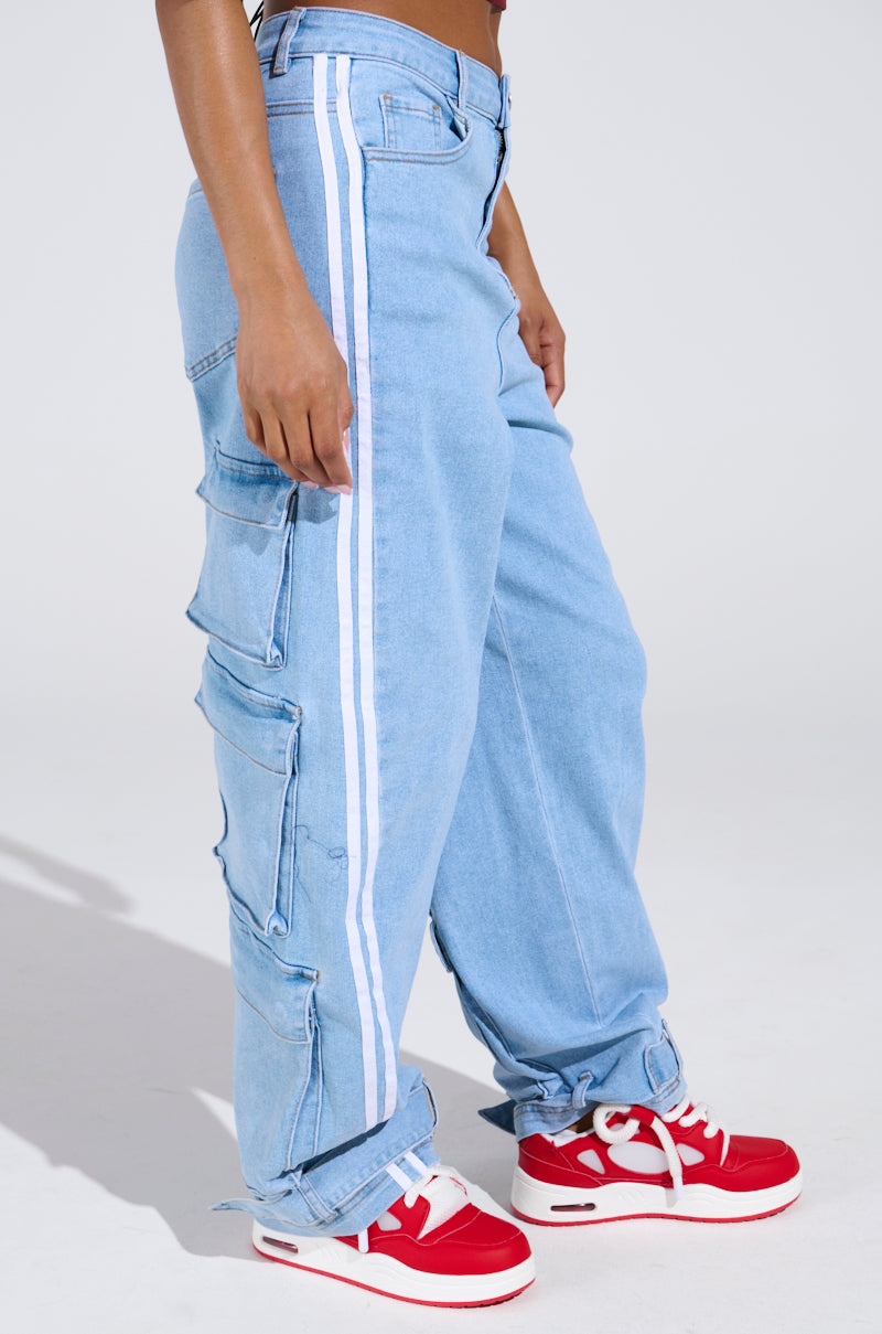 Someone stands against a plain light background wearing LEAVE IT ON THE TRACK RELAXED LEG DENIM PANTS, featuring white side stripes and large cargo pockets, paired with red-and-white sneakers.