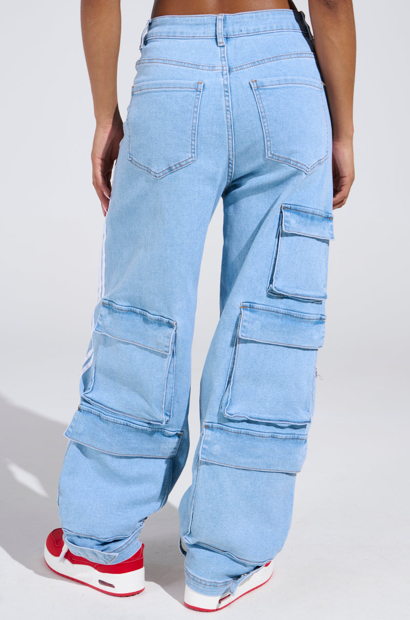 A person stands with their back to the camera, wearing LEAVE IT ON THE TRACK RELAXED LEG DENIM PANTS in light blue, featuring multiple large cargo pockets and a relaxed leg fit, paired with red and white sneakers.