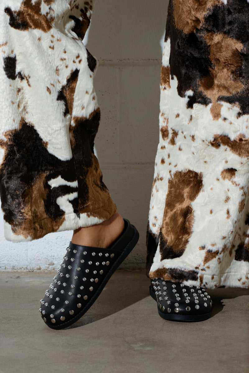 A person wearing cow-print pants is standing on a concrete floor, paired with AZALEA WANG TRAVERSE BLACK CLOGs. These stylish slip-ons are adorned with silver studs and feature comfort cushion insoles for a plush feel, blending fashion with practicality.