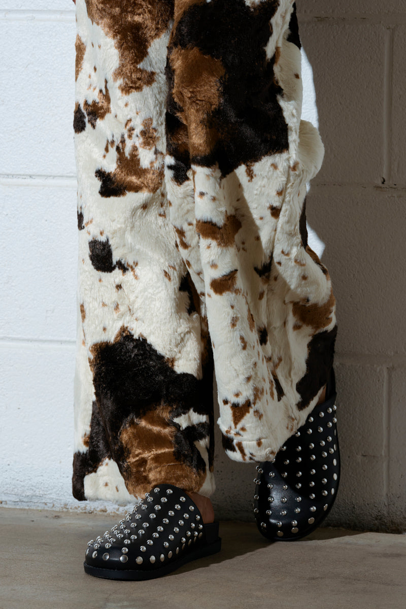 A person sports wide-leg pants in a brown and white cow pattern paired with AZALEA WANG TRAVERSE BLACK CLOG shoes featuring silver studs. They provide a perfect slip-on fit against a backdrop of a white brick wall.