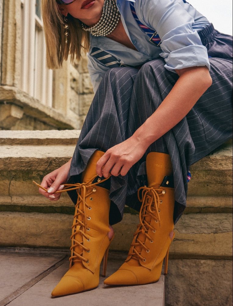 A woman sitting on stone steps wearing mustard yellow lace-up ankle boots with pointed toes and high stiletto heels, paired with a light blue shirt and dark pinstriped trousers.