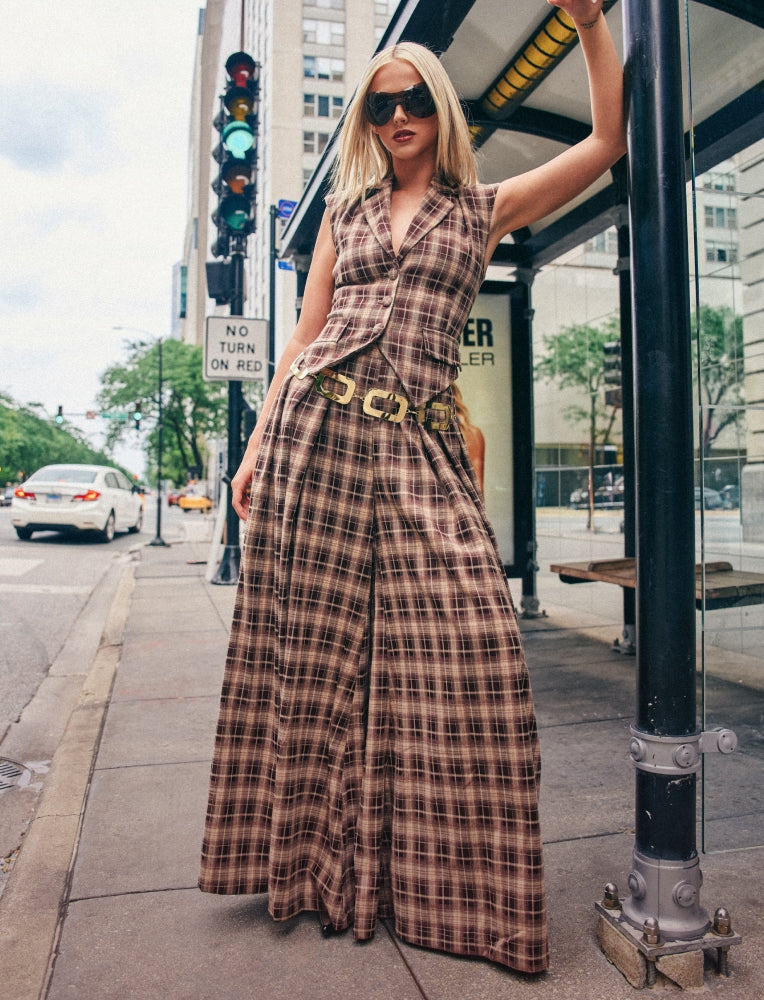 A woman wearing a brown and beige plaid wide-leg jumpsuit with a matching belt stands on a city sidewalk next to a bus stop shelter.