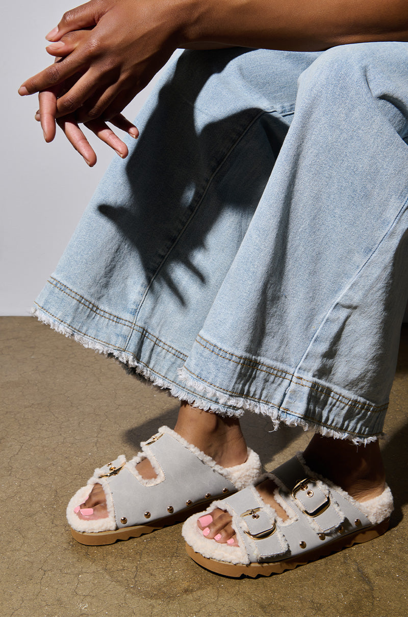 A person wearing light-colored denim jeans and AZALEA WANG TRESOR GREY FUZZY SANDALS featuring a flatform sole sits on a concrete floor, with the focus on the standout sandals and legs, while their hands rest calmly in their lap.