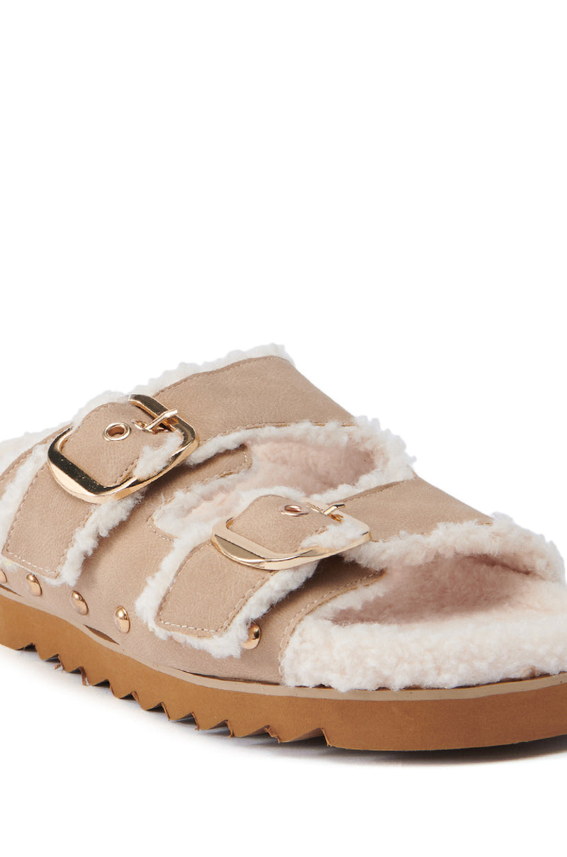 A close-up of the AZALEA WANG TRESOR TAN FUZZY SANDAL showcases its faux suede leather design, featuring two gold buckles and plush white lining, along with a brown sole with ridged texture for grip.
