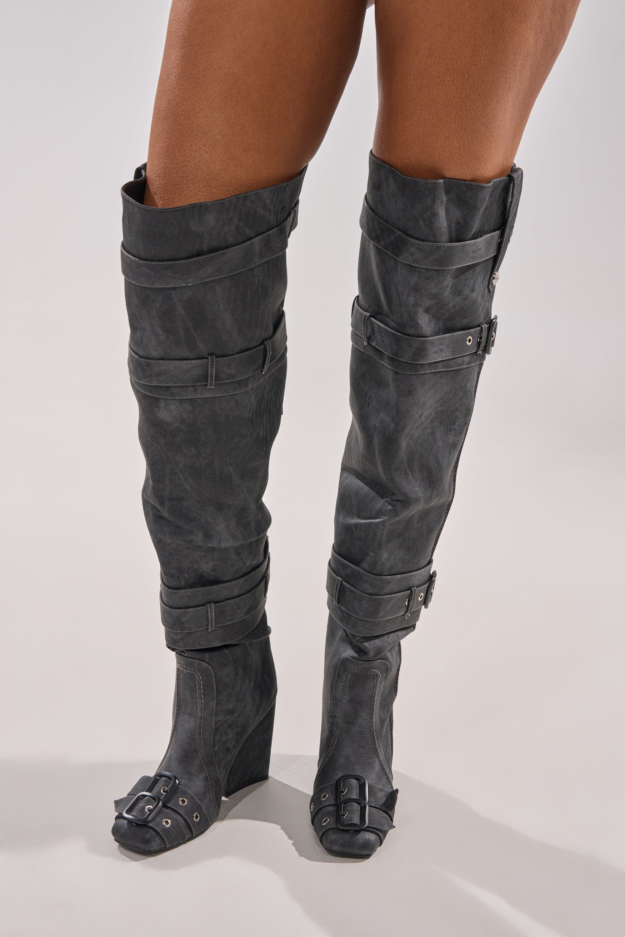 A person wears the AZALEA WANG TRILLION CHARCOAL CHUNKY THIGH HIGH BOOT, featuring black over-the-knee design with buckles and straps, standing on a plain light background to showcase its bold style.