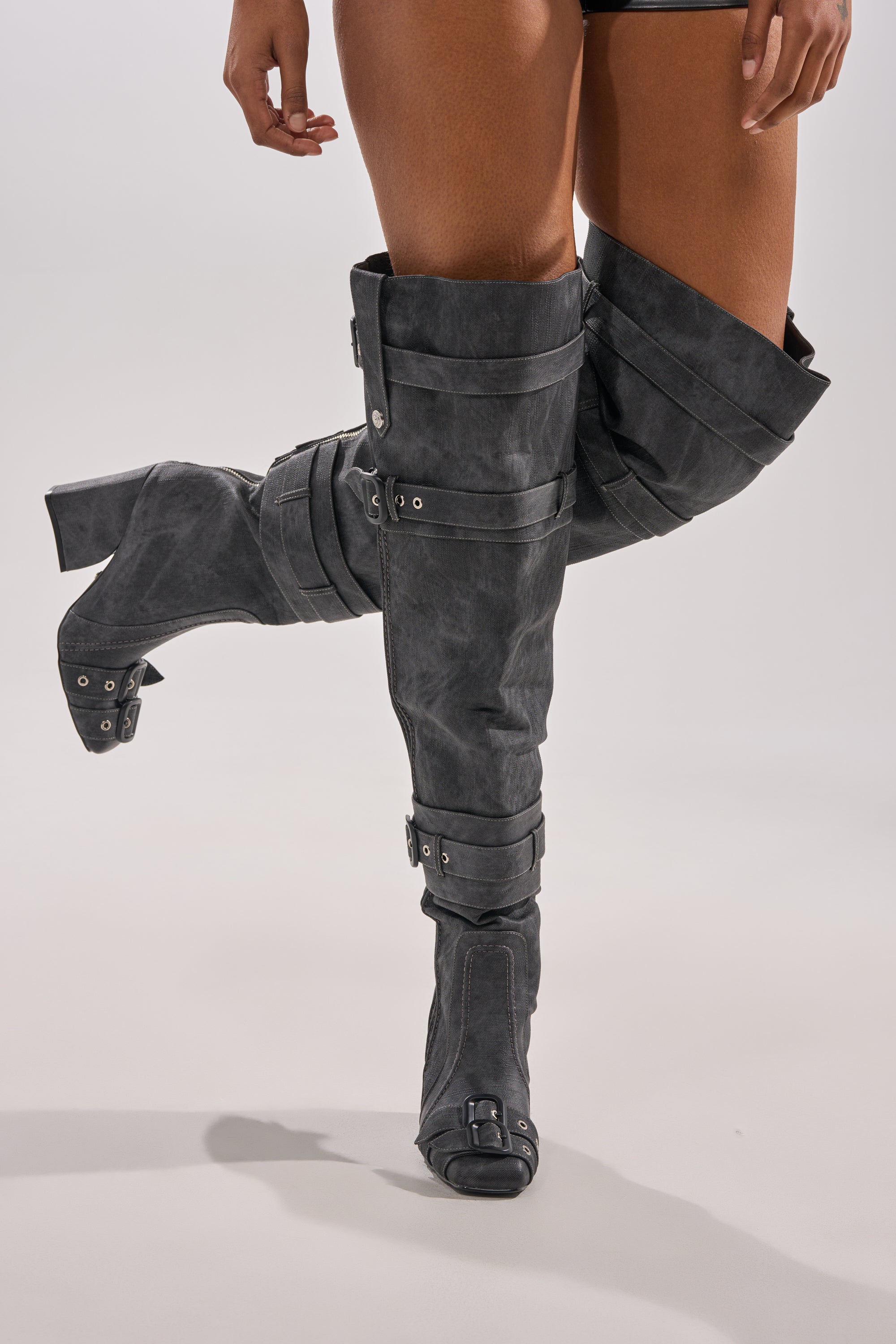 A person wears the AZALEA WANG TRILLION CHARCOAL CHUNKY THIGH HIGH BOOT, featuring black block heels with buckles and straps, standing with one leg crossed in front of the other against a neutral background.