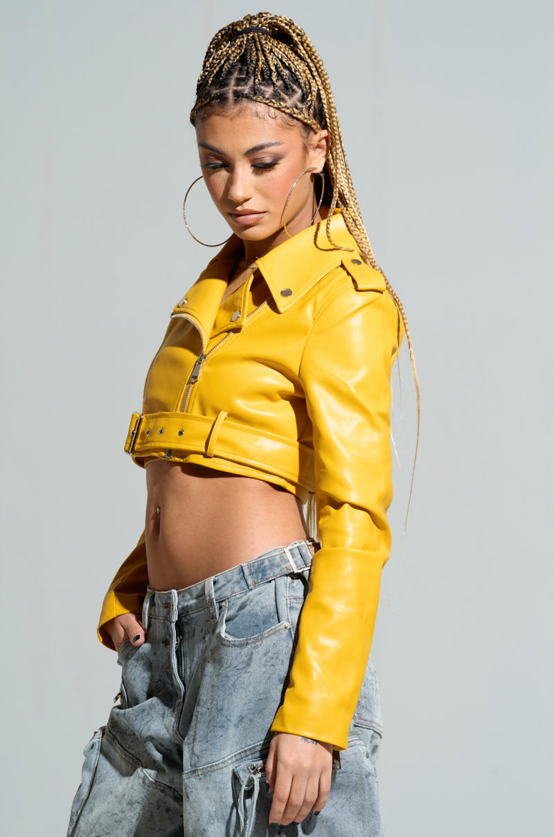 Person with braids wearing the TRIO ESSENTIAL CROPPED MOTO IN YELLOW and denim jeans, posing with a serious expression. They have large hoop earrings and an adjustable waist belt, standing against a plain backdrop.