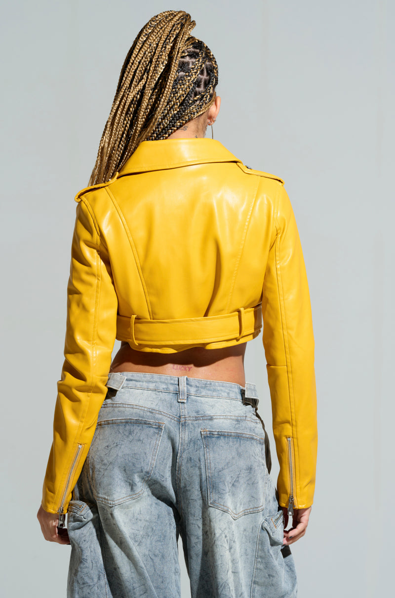A person with braided hair faces away, showcasing the TRIO ESSENTIAL CROPPED MOTO IN YELLOW, a bright faux leather jacket. It's paired with faded blue jeans and features an adjustable waist belt. The backdrop is a light gray wall.