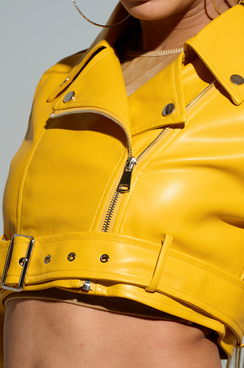 Close-up of a person wearing the TRIO ESSENTIAL CROPPED MOTO IN YELLOW, a striking faux leather jacket adorned with silver buttons and a zipper. This cropped moto jacket boasts a wide collar and an adjustable waist belt, set against a light neutral background.