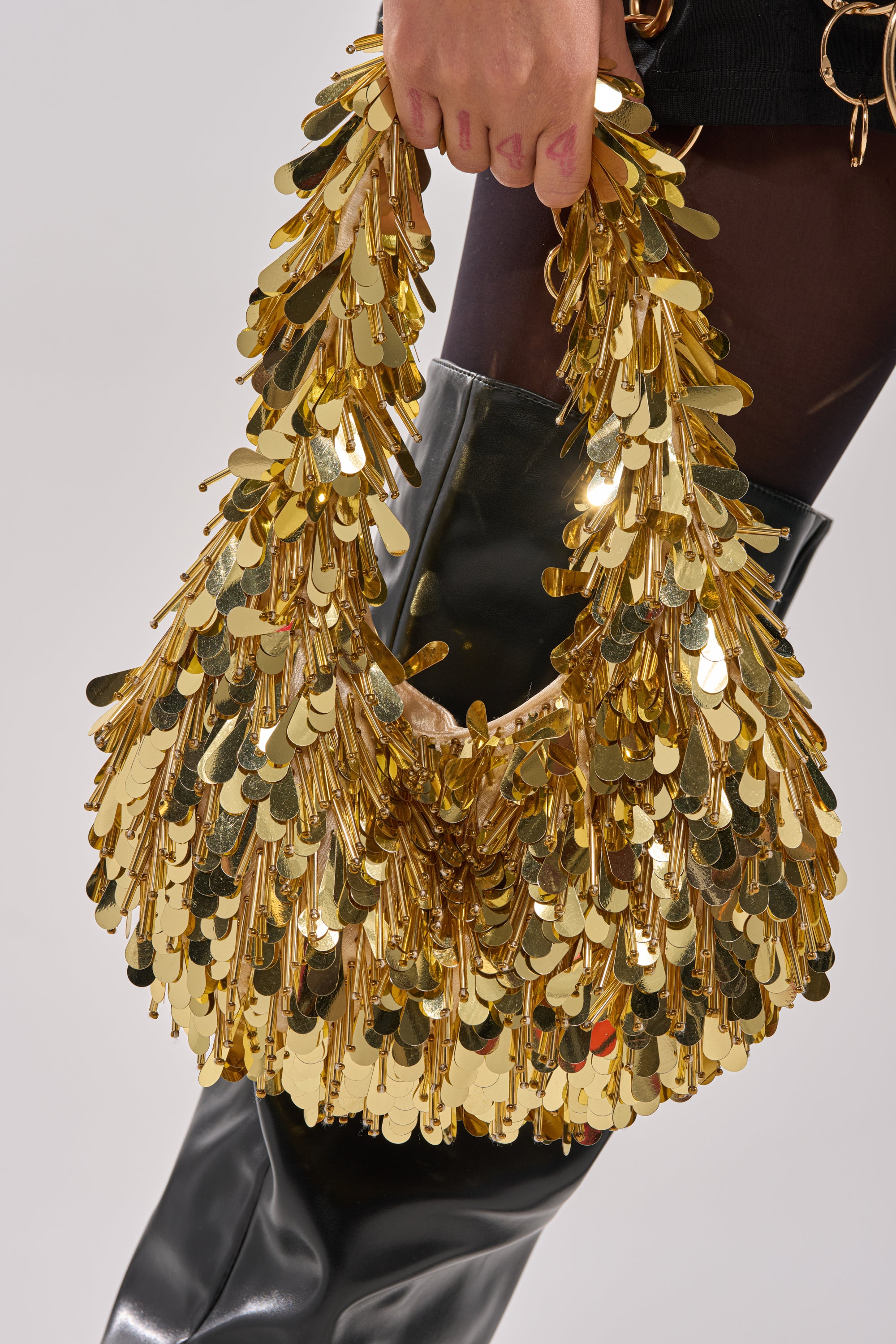 A person holds THE NOSTALGIA SEQUIN PURSE IN GOLD, featuring large, shiny gold overlapping discs and beaded details, paired with black boots and sheer black tights.