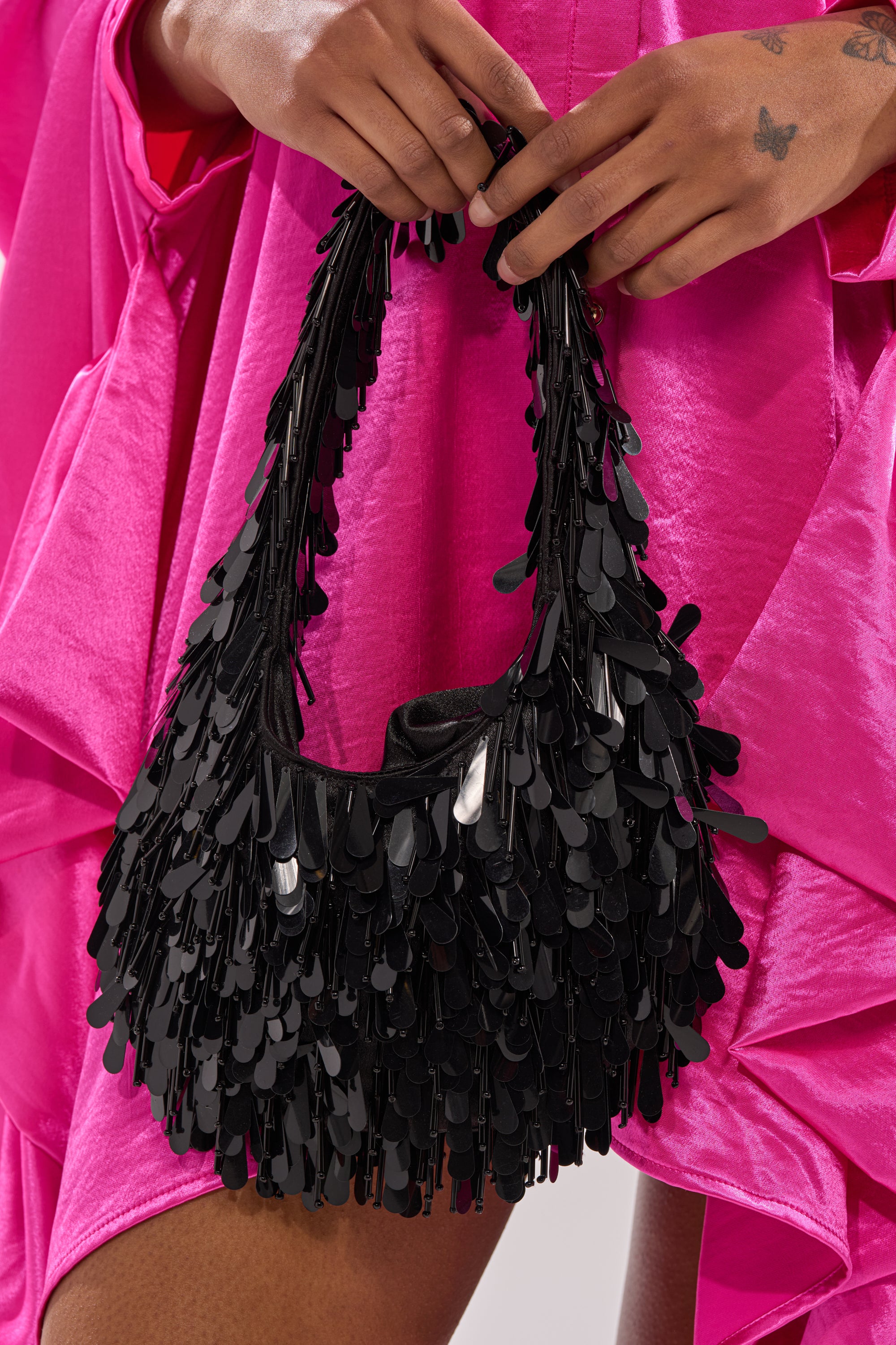 A person with manicured nails and small hand tattoos holds THE NOSTALGIA SEQUIN PURSE IN BLACK—a black fringe shoulder bag decorated with shiny, teardrop-shaped sequins—while wearing a bright pink textured dress.