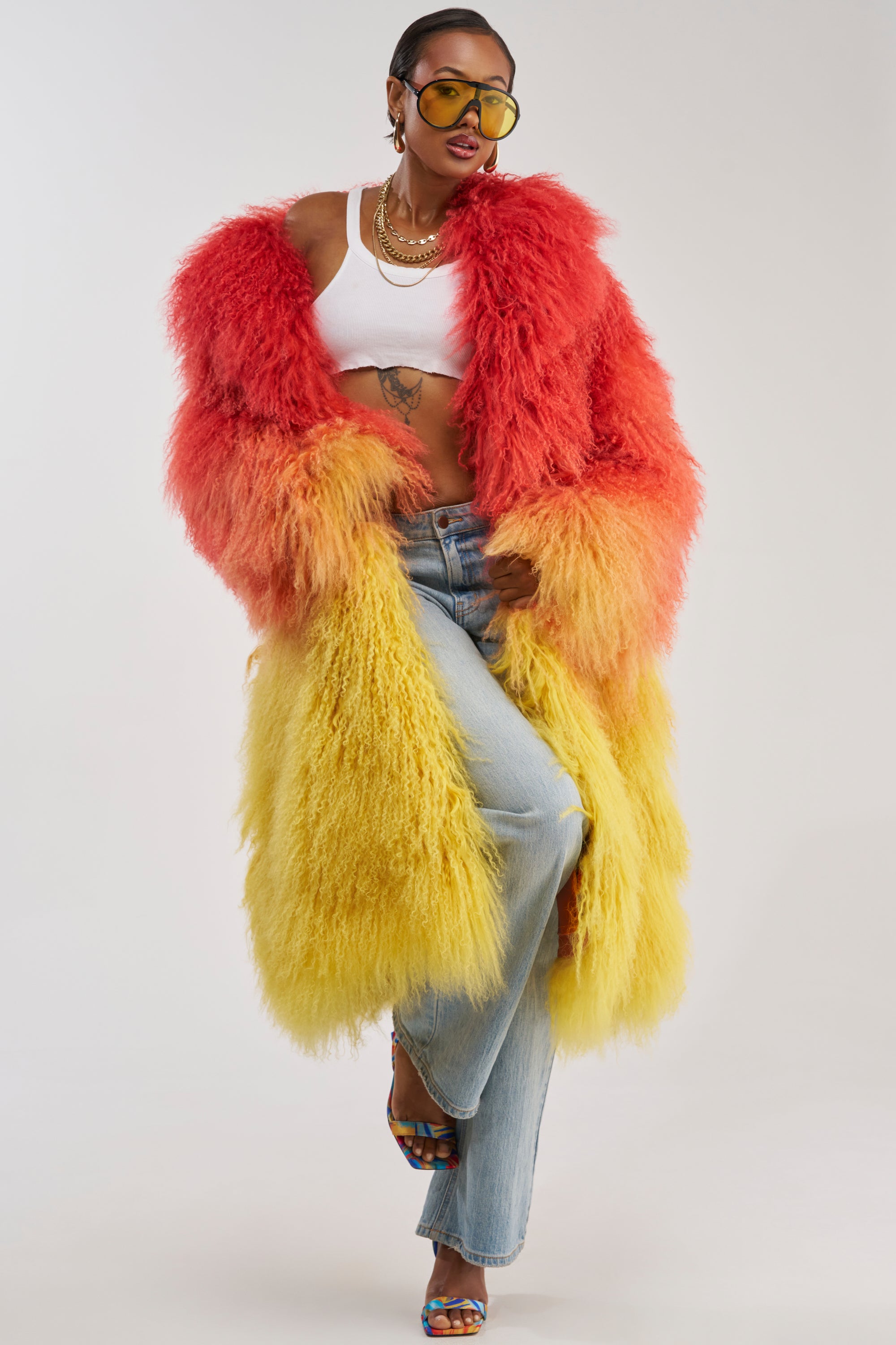 Wearing the TROPEZ GENUINE MONGOLIAN FUR COAT—a striking fluffy red and yellow ombre longline coat—paired with a white crop top, light blue jeans, layered necklaces, yellow sunglasses, and multicolored heels, the person poses confidently against a plain background.