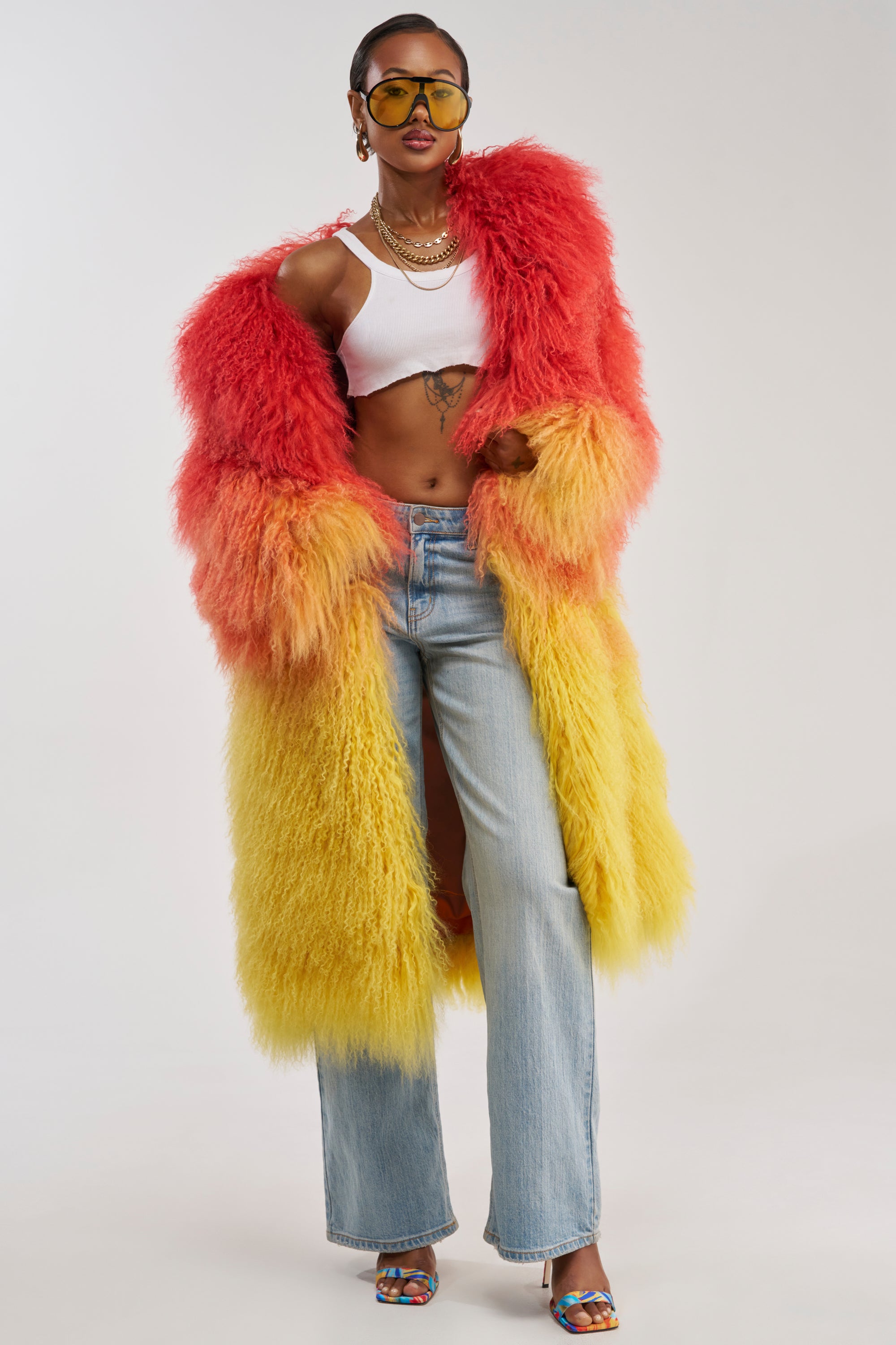 Wearing the TROPEZ GENUINE MONGOLIAN FUR COAT, featuring a bold ombre design in red and yellow hues with a luxurious fuzzy Mongolian fur texture, this individual pairs it with a white crop top, blue jeans, sunglasses, and colorful heels while posing confidently against a plain background.
