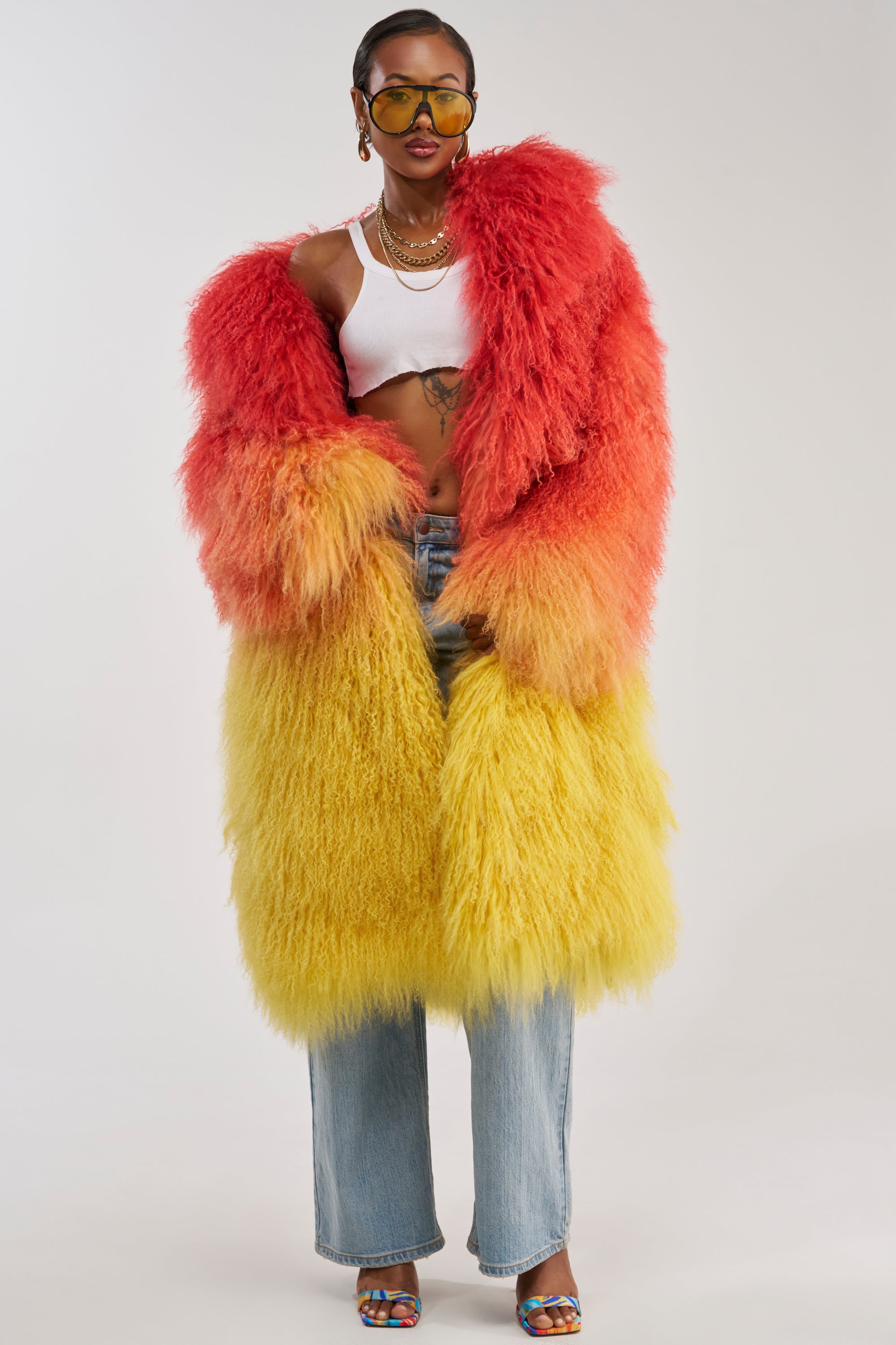 Wearing the TROPEZ GENUINE MONGOLIAN FUR COAT in a striking red and yellow ombre, paired with a white crop top, blue jeans, and sunglasses, a person stands confidently against a plain background.
