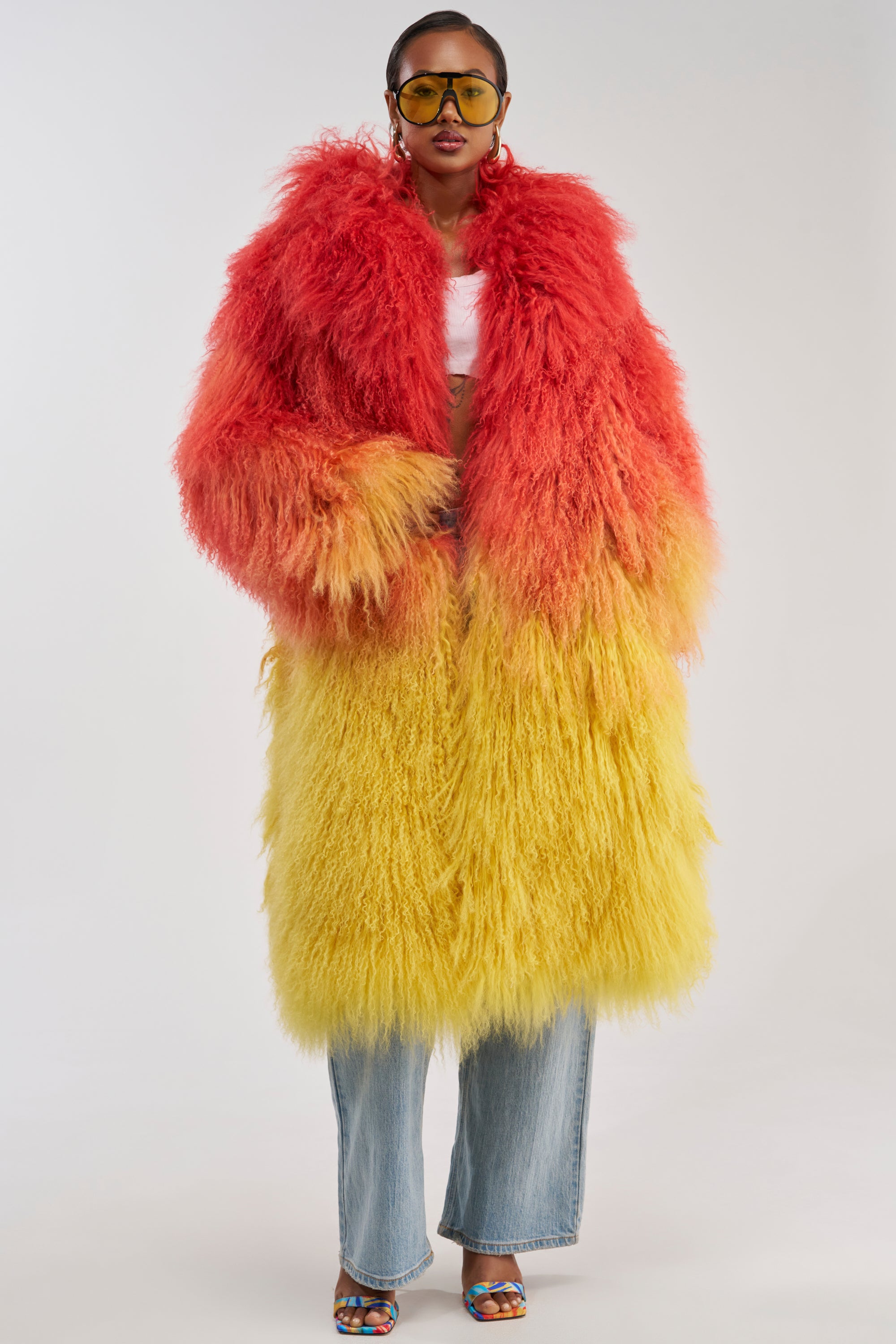 Wearing the TROPEZ GENUINE MONGOLIAN FUR COAT with a red and yellow gradient, paired with blue jeans, colorful heels, a white top, and yellow sunglasses, a person stands against a plain background.