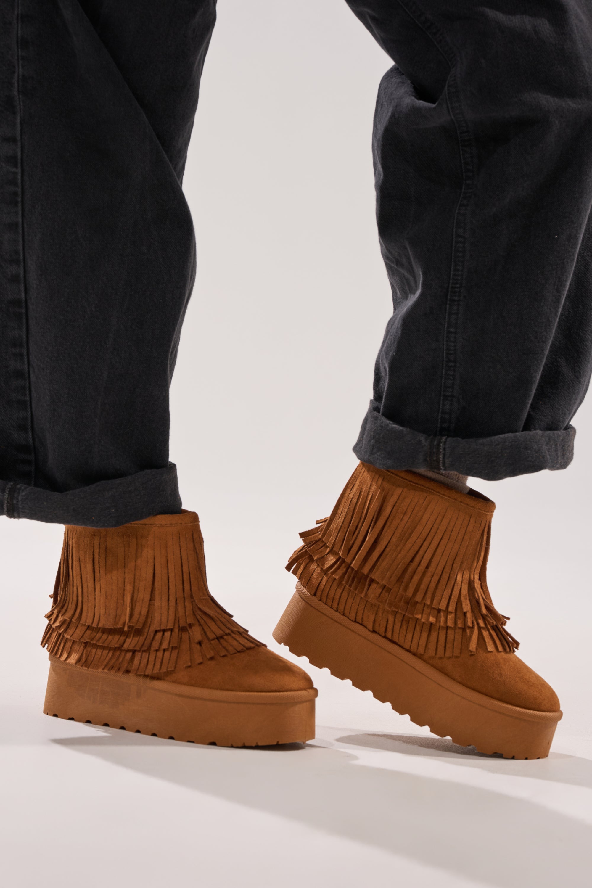 Wearing cuffed black jeans and AKIRA BROWN FAUX SUEDE FRINGE BOOTIE with a flatform sole, a person stands on a light background.
