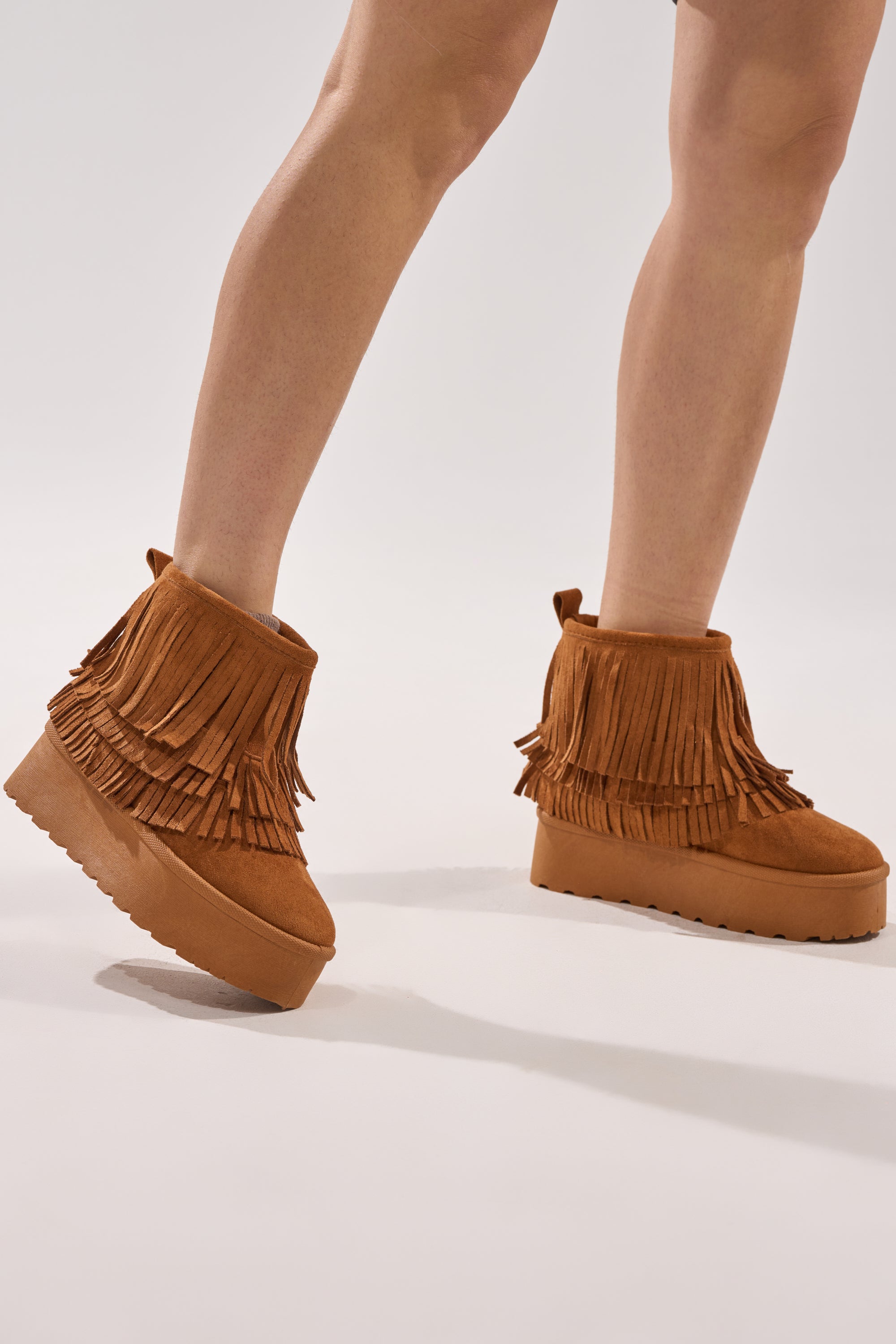 A person models the AKIRA BROWN FAUX SUEDE FRINGE BOOTIE with a flatform sole, standing on a light background. Only their legs and feet are visible.