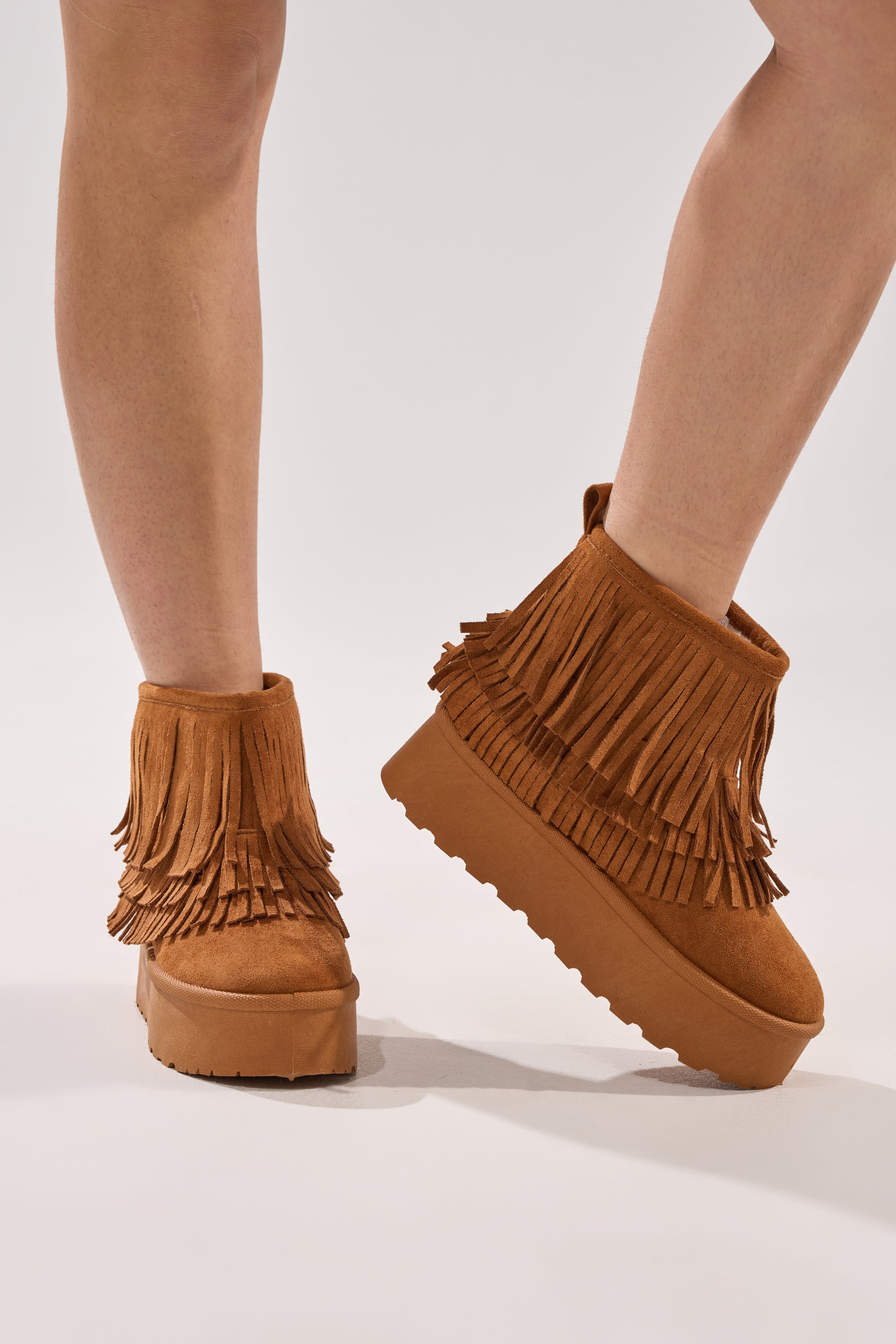 Someone stands on a light surface with legs slightly crossed, wearing the AKIRA BROWN FAUX SUEDE FRINGE BOOTIE featuring a flatform sole.